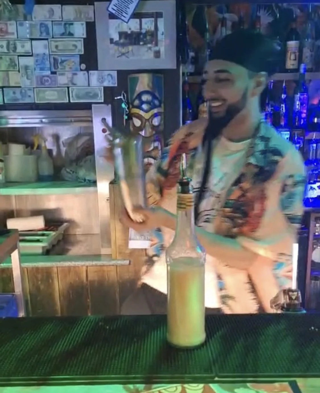 A bartender with a beard and a colorful Hawaiian shirt stands behind the bar, smiling and holding a bottle with a straw. The background has various liquor bottles and decorative items, including dollar bills taped to the wall.