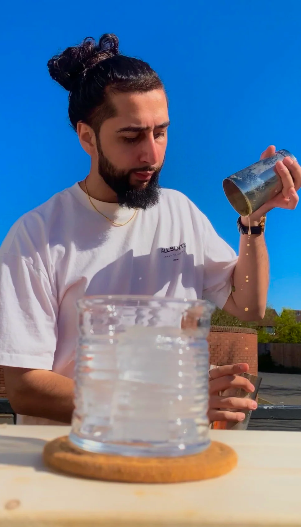 A young man with dark hair in a top knot, beard, wearing a white t-shirt with text, is pouring liquid from a metallic cup into a glass container on a wooden surface outdoors on a sunny day.