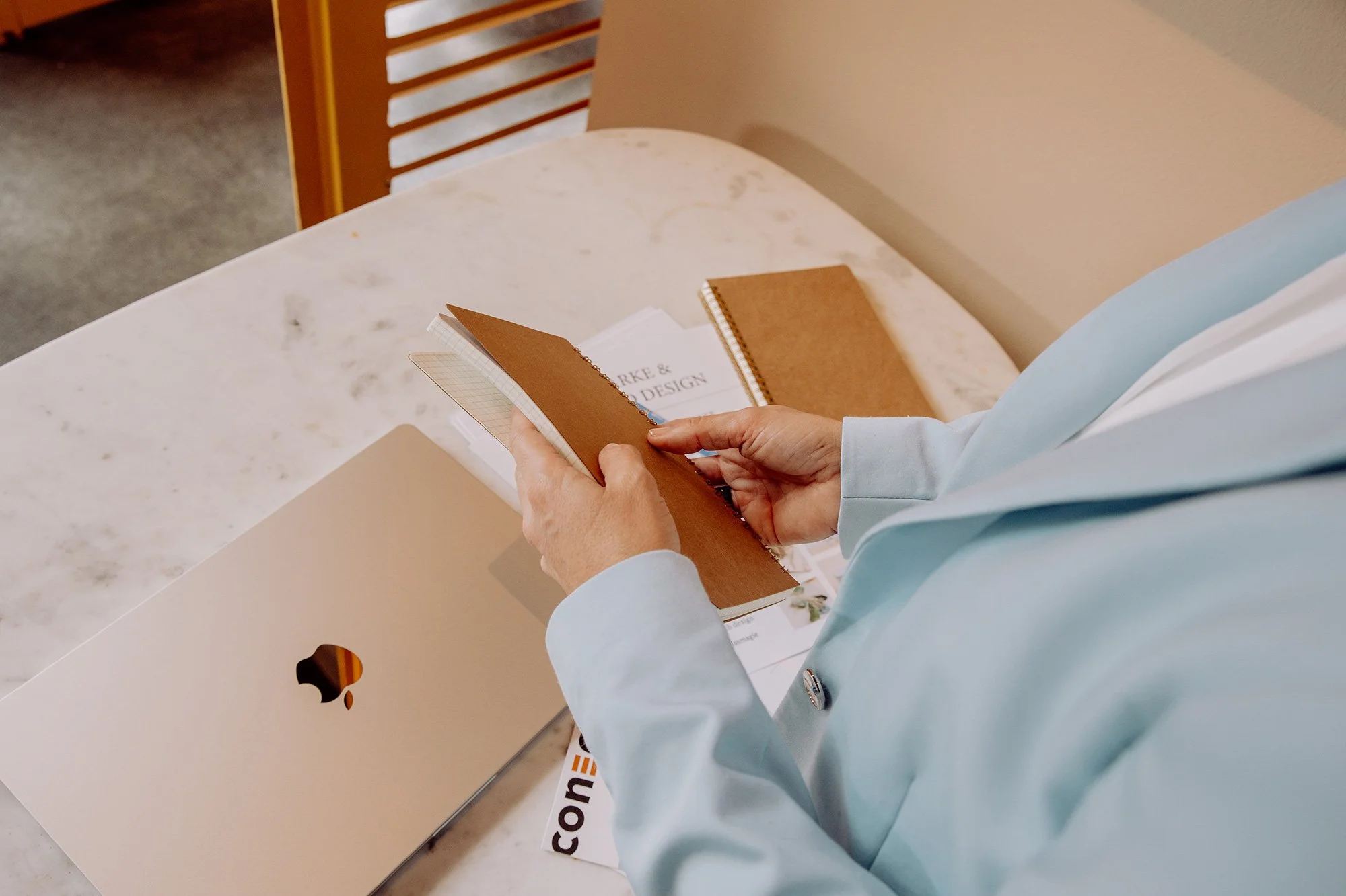 Person liest in einem Notizbuch auf einem Tisch mit weiteren Notizbüchern, einem Laptop und Unterlagen, in einer hellen Umgebung.