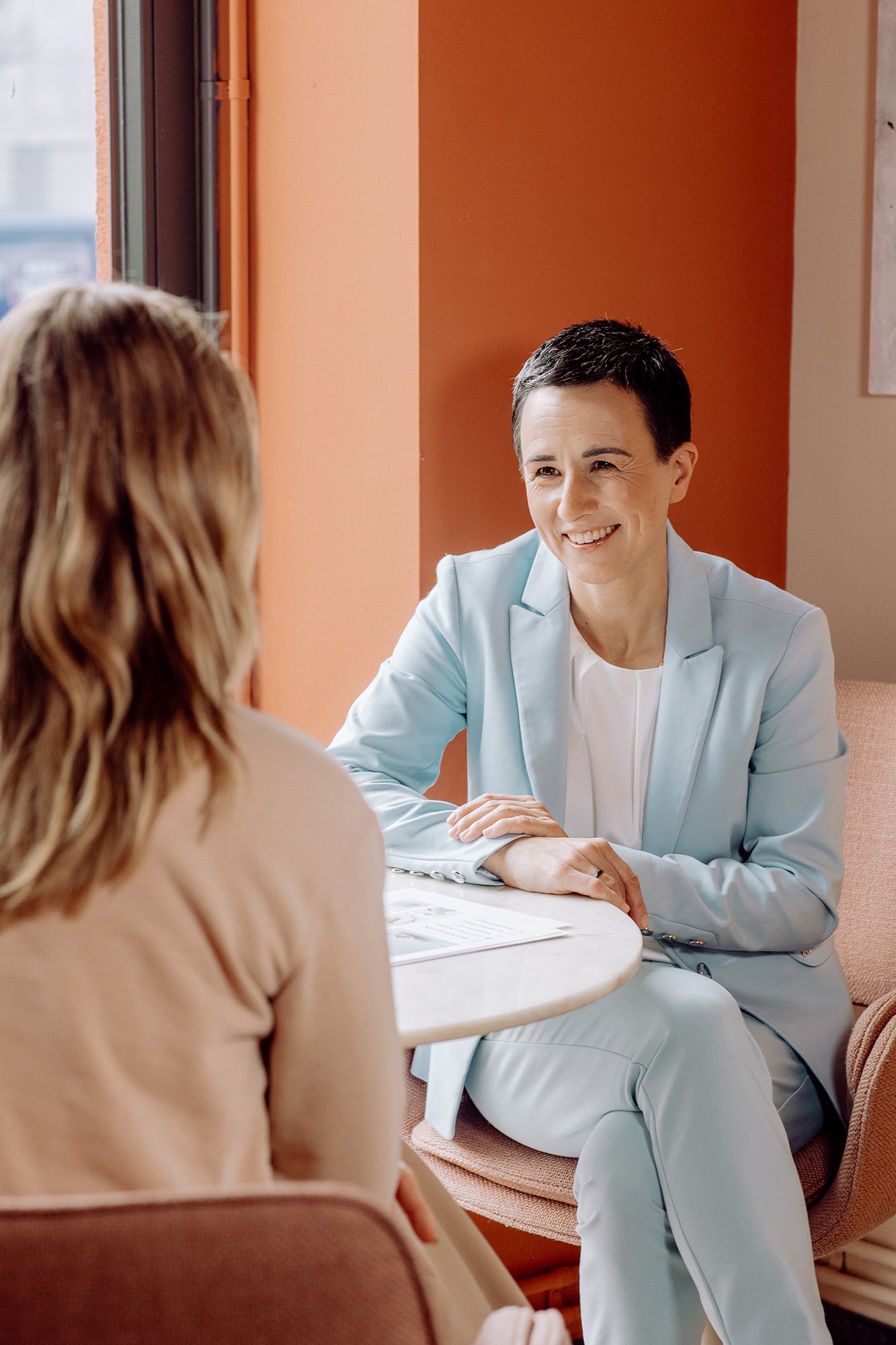 Zwei Frauen sitzen sich an einem Tisch bei einem Gespräch in einem hell erleuchteten Raum mit orangefarbenen Wänden. Die Frau auf der rechten Seite trägt ein hellblaues Jackett und lacht freundlich.