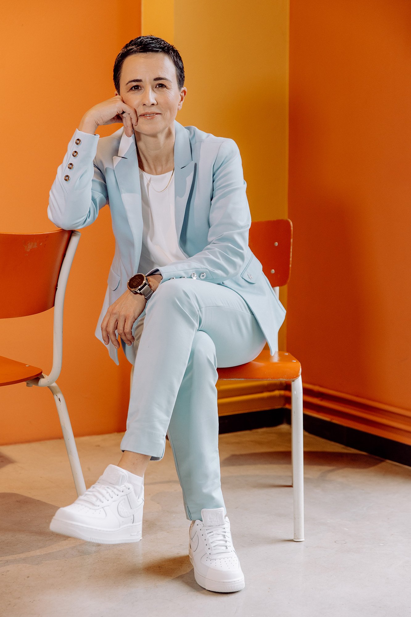 Frau in heller Anzug sitzt auf einem orangefarbenen Stuhl vor einer orangefarbenen Wand.