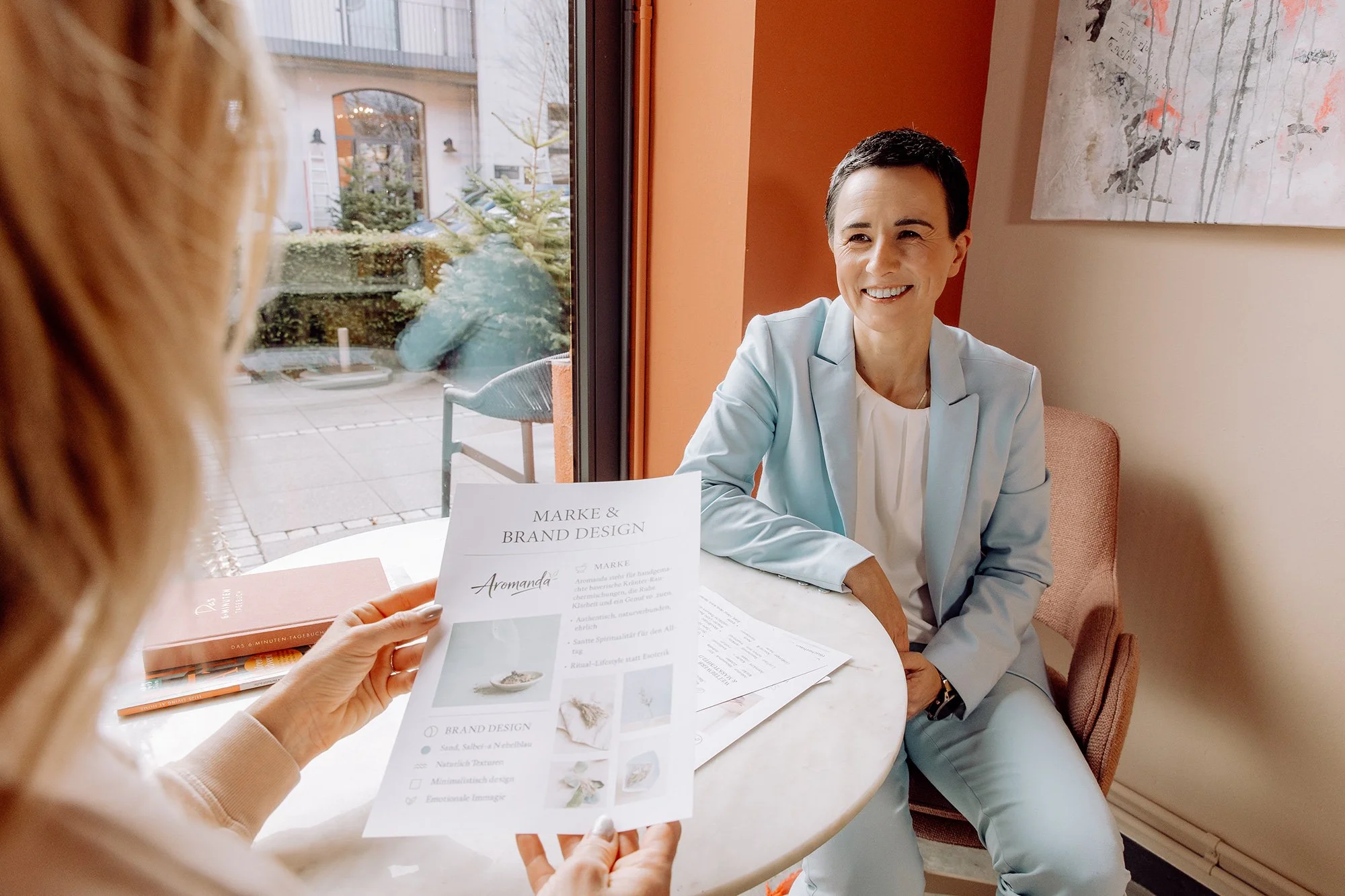 Zwei Frauen im Gespräch in einem Café, eine hält einen Flyer mit Text und Bildern, die andere sitzt lächelnd am Tisch, draußen sind Bäume und Gebäude sichtbar.
