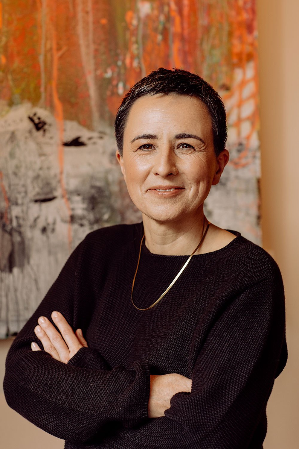 Eine lächelnde Frau mit kurzen dunklen Haaren, trägt eine schwarze Strickjacke und eine goldene Halskette, vor einem bunten Kunstwerk