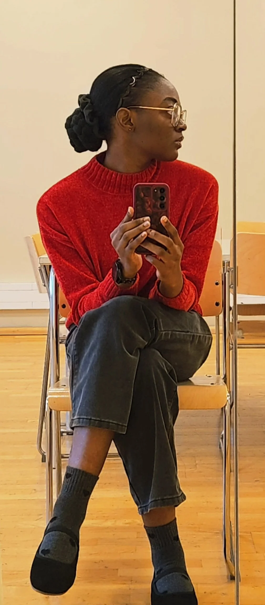 Person sitting on a chair taking a mirror selfie, wearing a red sweater, dark pants, and colorful socks, with glasses and braided hair, in a room with wooden floors.