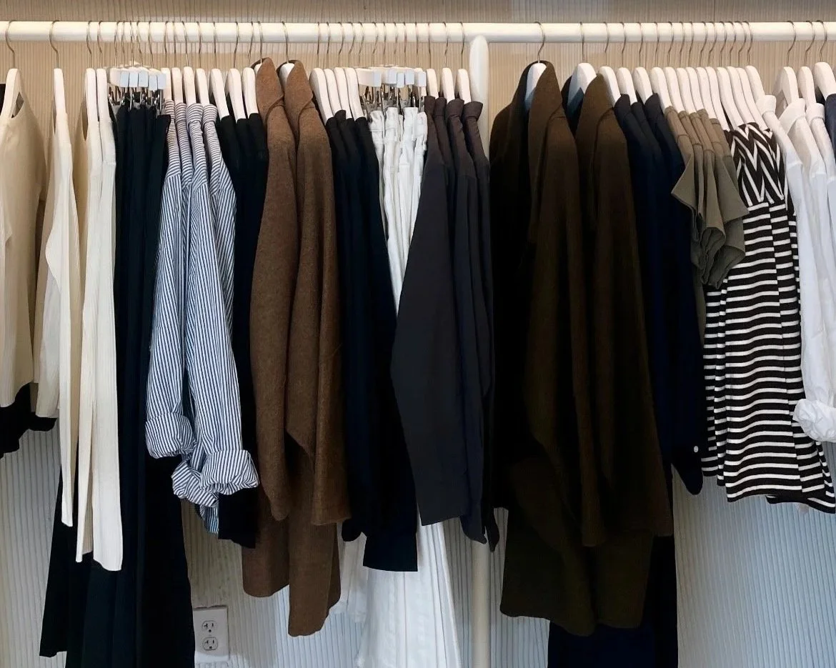 A clothing rack with various men's and women's garments including shirts, pants, and jackets in colors like white, black, brown, navy, and striped patterns.