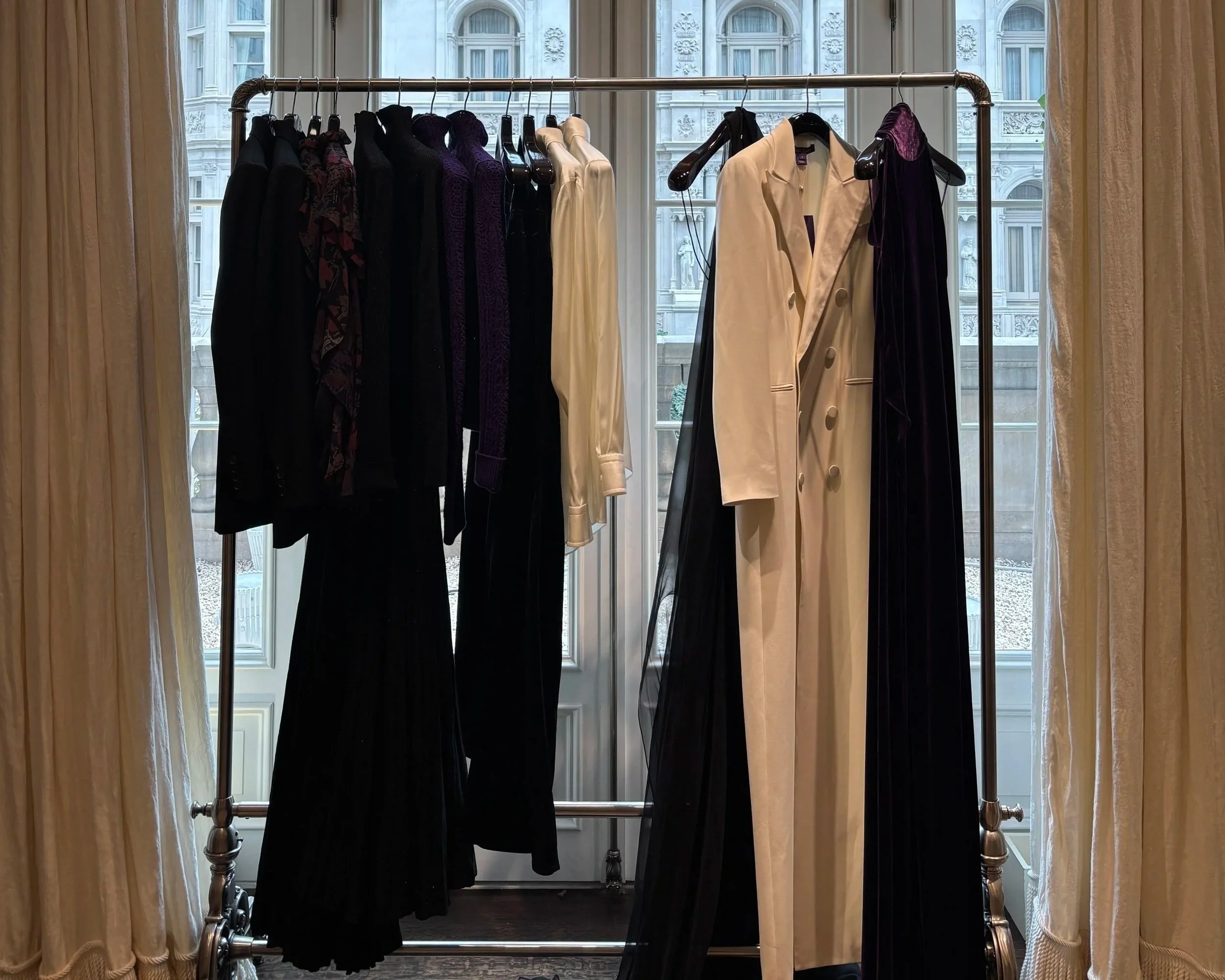 Clothing rack with dresses and coats inside a room with large window and curtains, city buildings in the background.