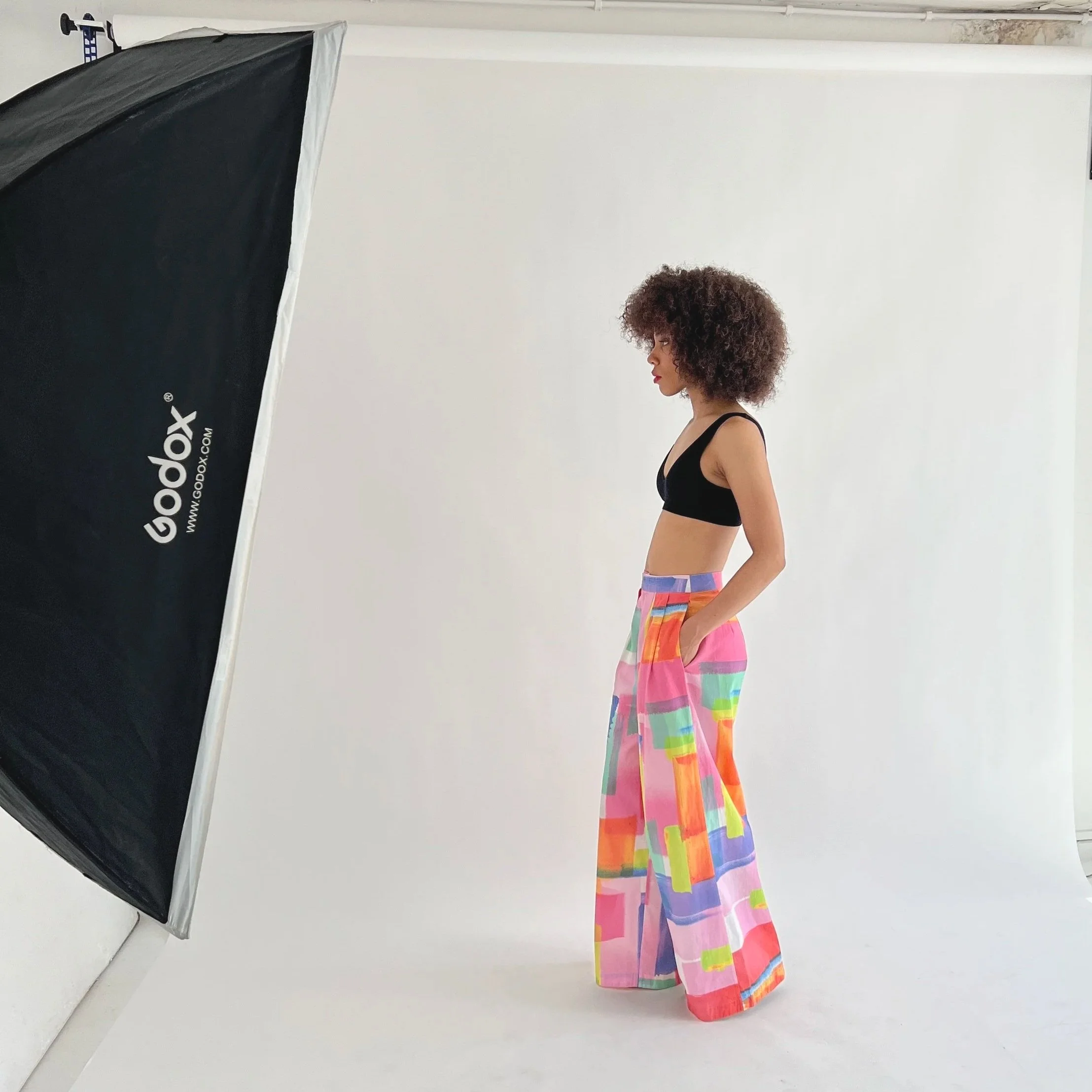 A woman with curly hair stands in a photography studio, wearing a black crop top and colorful wide-leg pants with a geometric pattern, while facing sideways near a white backdrop and a large Godox photography light.