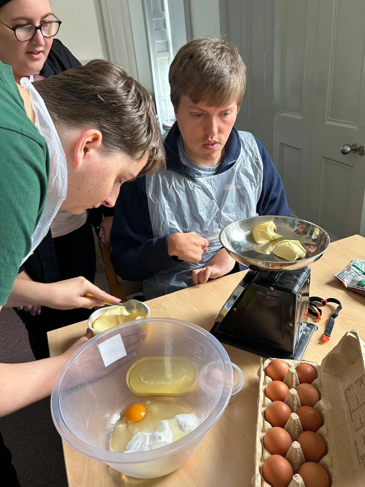 A group of people baking, with a young man in a blue sweater and apron, weighing butter on a scale, eggs in a carton, and a bowl with cracked eggs, butter, and measuring cups.
