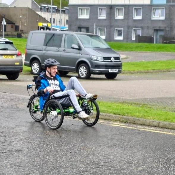 Person riding a handcycle on a city street, wearing a helmet and a blue jacket, with parked cars and gray buildings in the background.