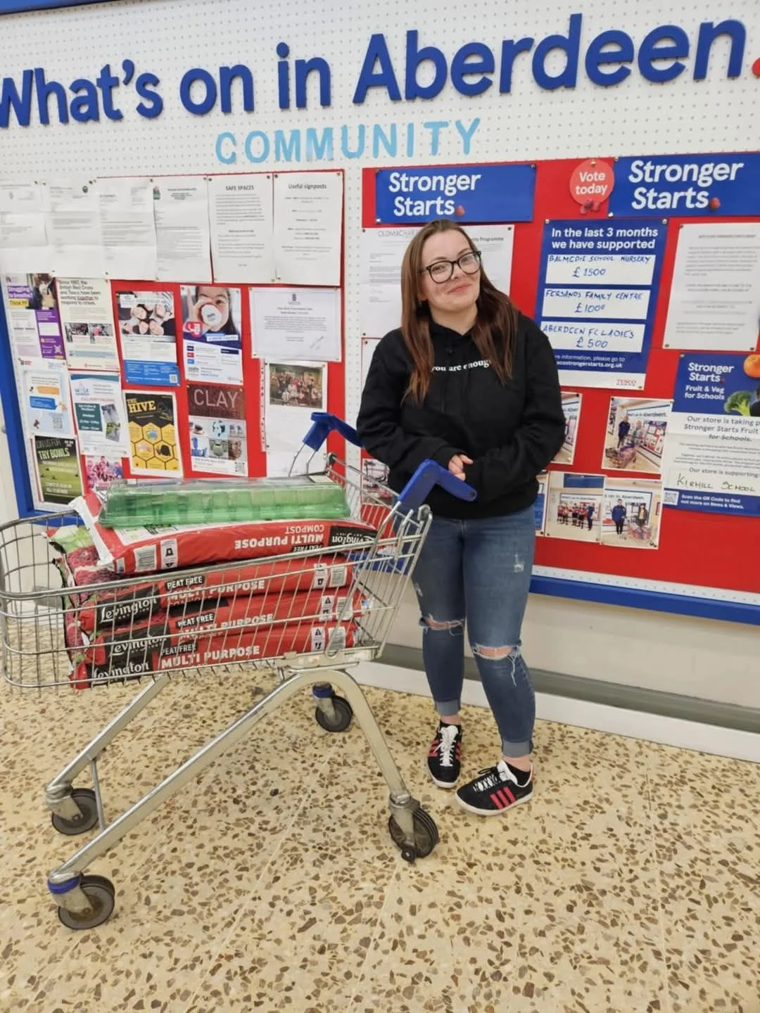 A young woman with long brown hair, glasses, wearing a black hoodie, ripped jeans, and black sneakers with red and white stripes, standing inside a store next to a shopping cart filled with gardening supplies, in front of a community bulletin board.