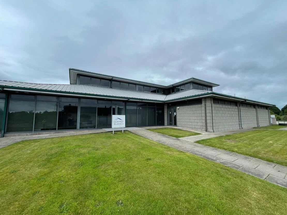 A modern, single-story building with glass walls, a metal roof, and a small concrete pathway leading to the entrance, surrounded by green grass and an overcast sky.