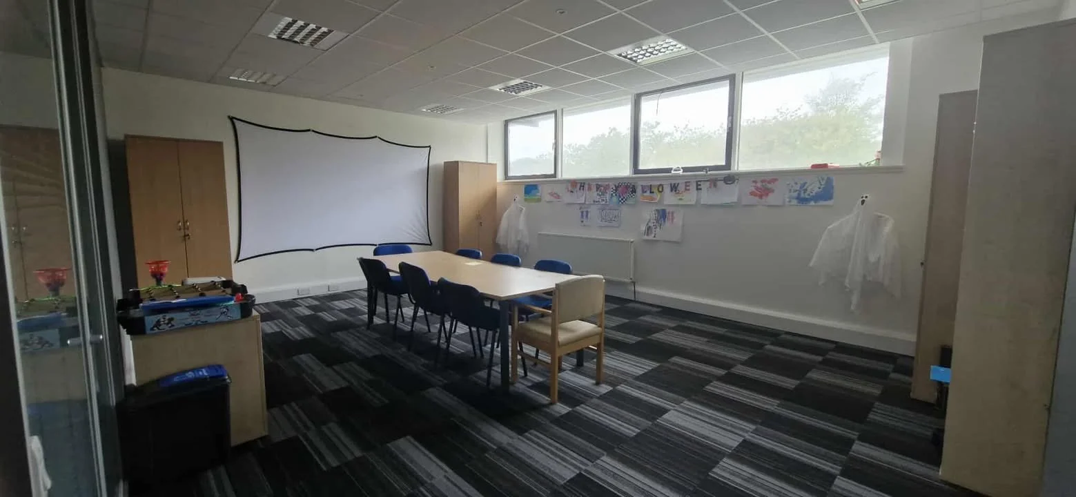Empty classroom with a large table surrounded by chairs, a white screen on the wall, and child's artwork including Halloween-themed decorations and drawings displayed along the wall. Natural light coming from windows.