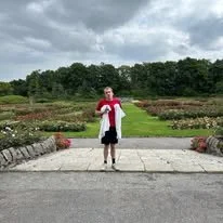 Person standing on a sidewalk in front of a landscaped garden with trees and cloudy sky