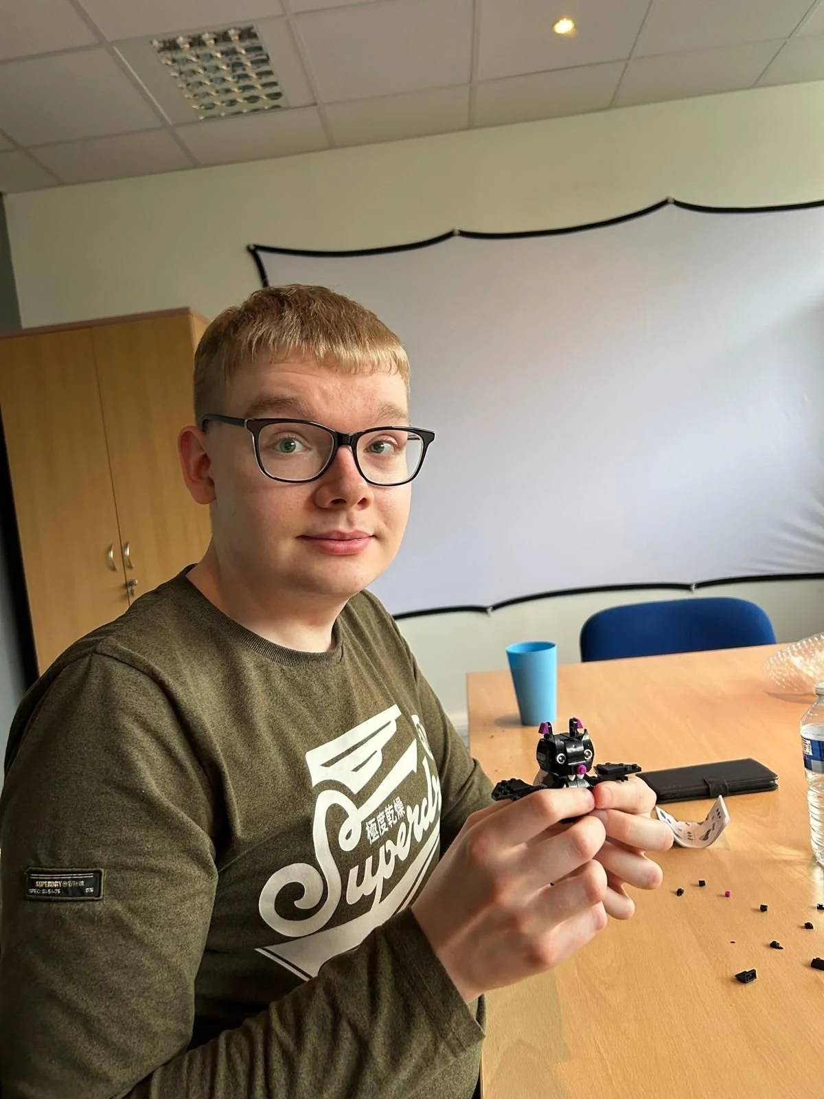 A young man with glasses sitting at a wooden table, holding a small assembled robot in his hands. The table has a blue cup, a water bottle, and scattered LEGO pieces. Behind him is a white projection screen and a wooden cabinet in a room with a dropp