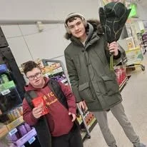Two young men shopping in a store, one holding a bouquet of flowers and giving a thumbs-up, both wearing jackets and glasses.