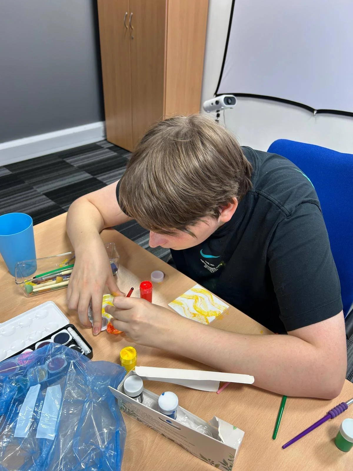 A young boy sitting at a table, engaged in a painting or craft project with various paints, brushes, and supplies around him.