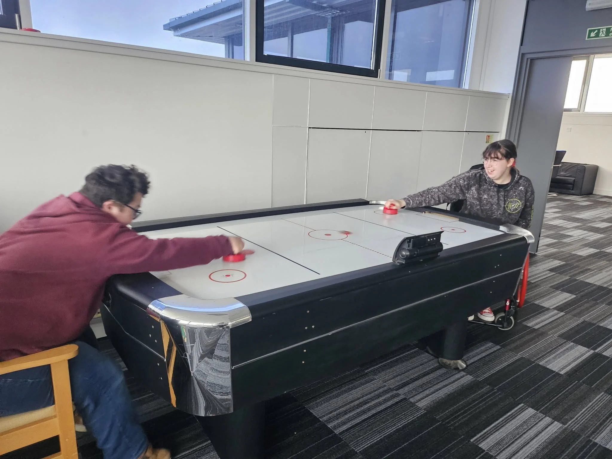 Two people playing air hockey indoors, one on the left has dark hair and glasses, wearing a maroon hoodie, and the other on the right with glasses, wearing a dark hoodie, smiling.
