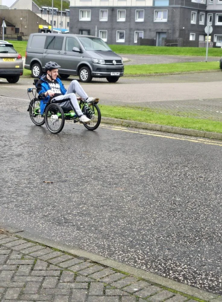 A person riding a recumbent tricycle on a wet street in an urban area, with parked cars and residential buildings in the background.