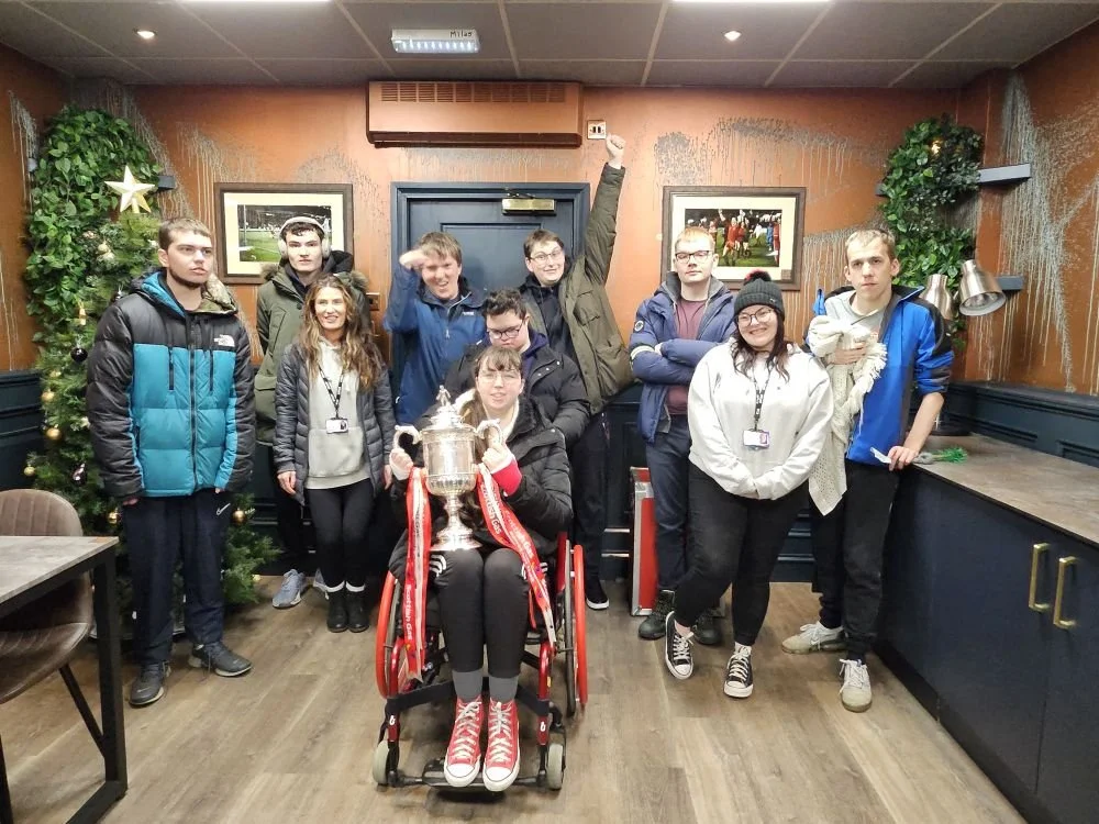 Group of ten young people, including a girl in a wheelchair holding a trophy, celebrating indoors with holiday decorations, including a Christmas tree, framed photos, and festive ornaments.