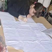 A woman sitting at a table, writing on a large piece of paper with a pen