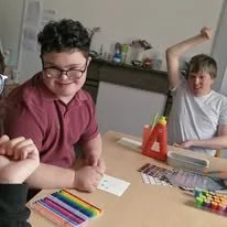 Two boys sitting at a table, one smiling and the other raising his arm behind his head, with colorful markers on the table.