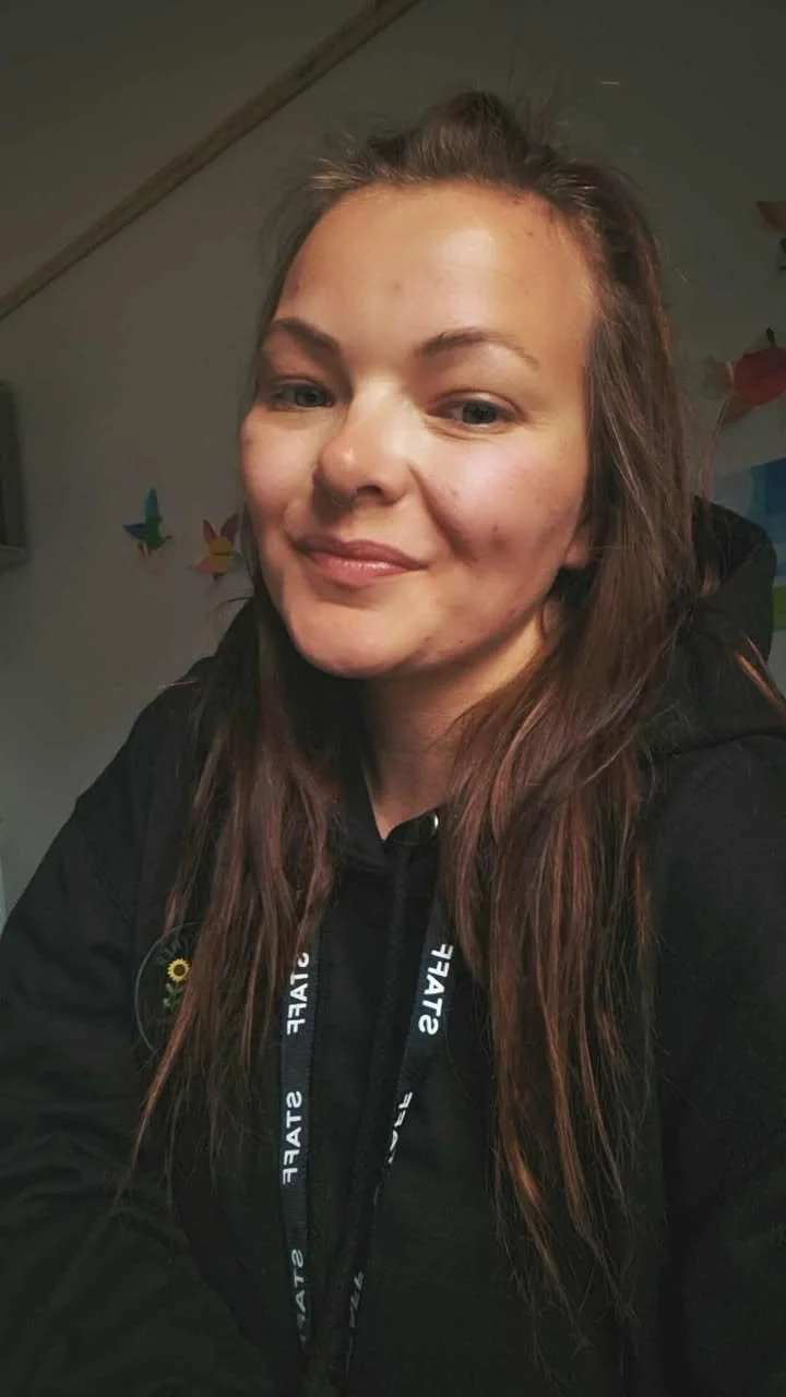 A woman with long brown hair, smiling, wearing a black hoodie with a lanyard around her neck, indoors with butterfly decorations on the wall in the background.