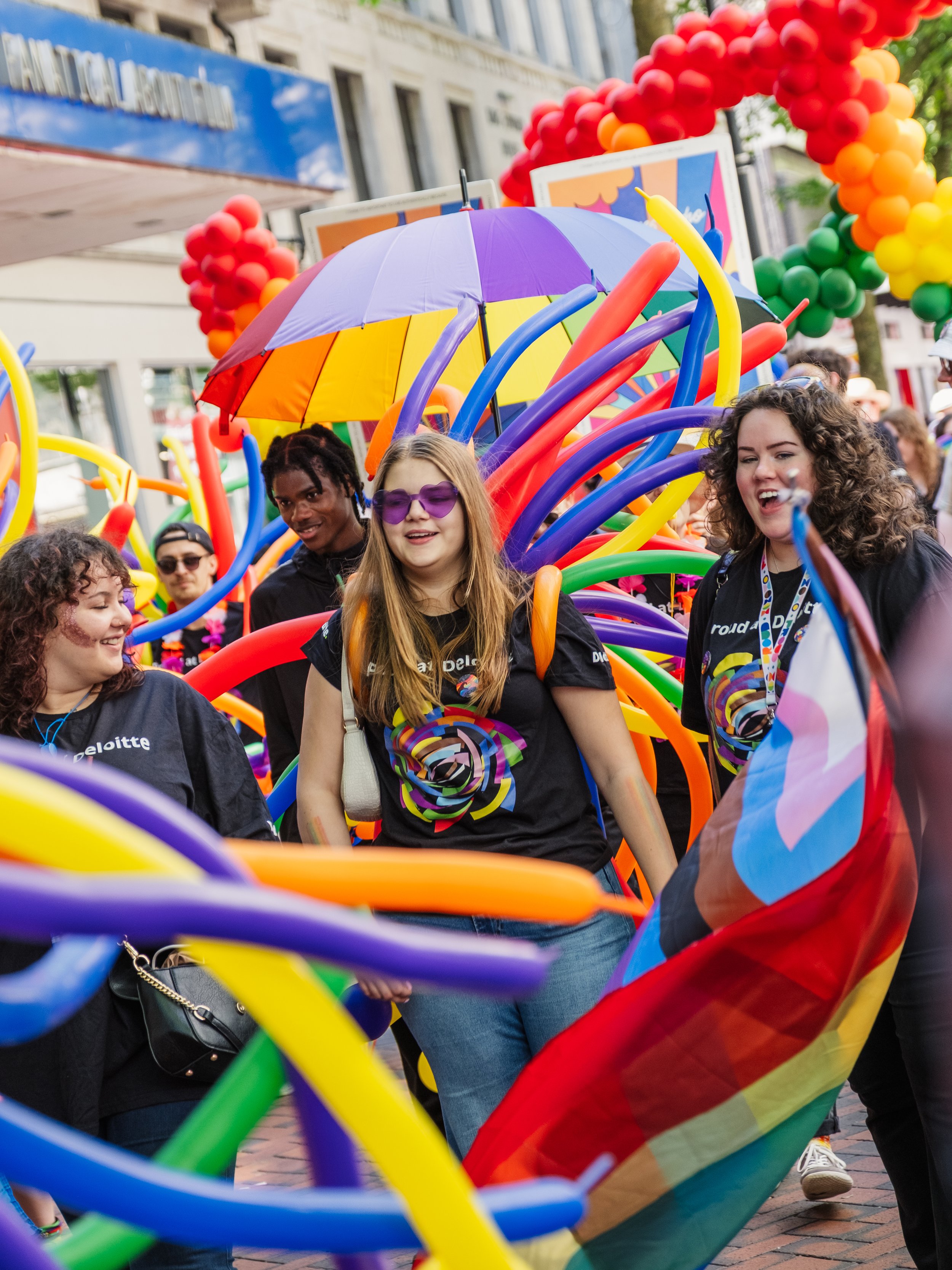 People participating in a pride parade with rainbow-colored balloons, flags, and accessories, walking down a city street.