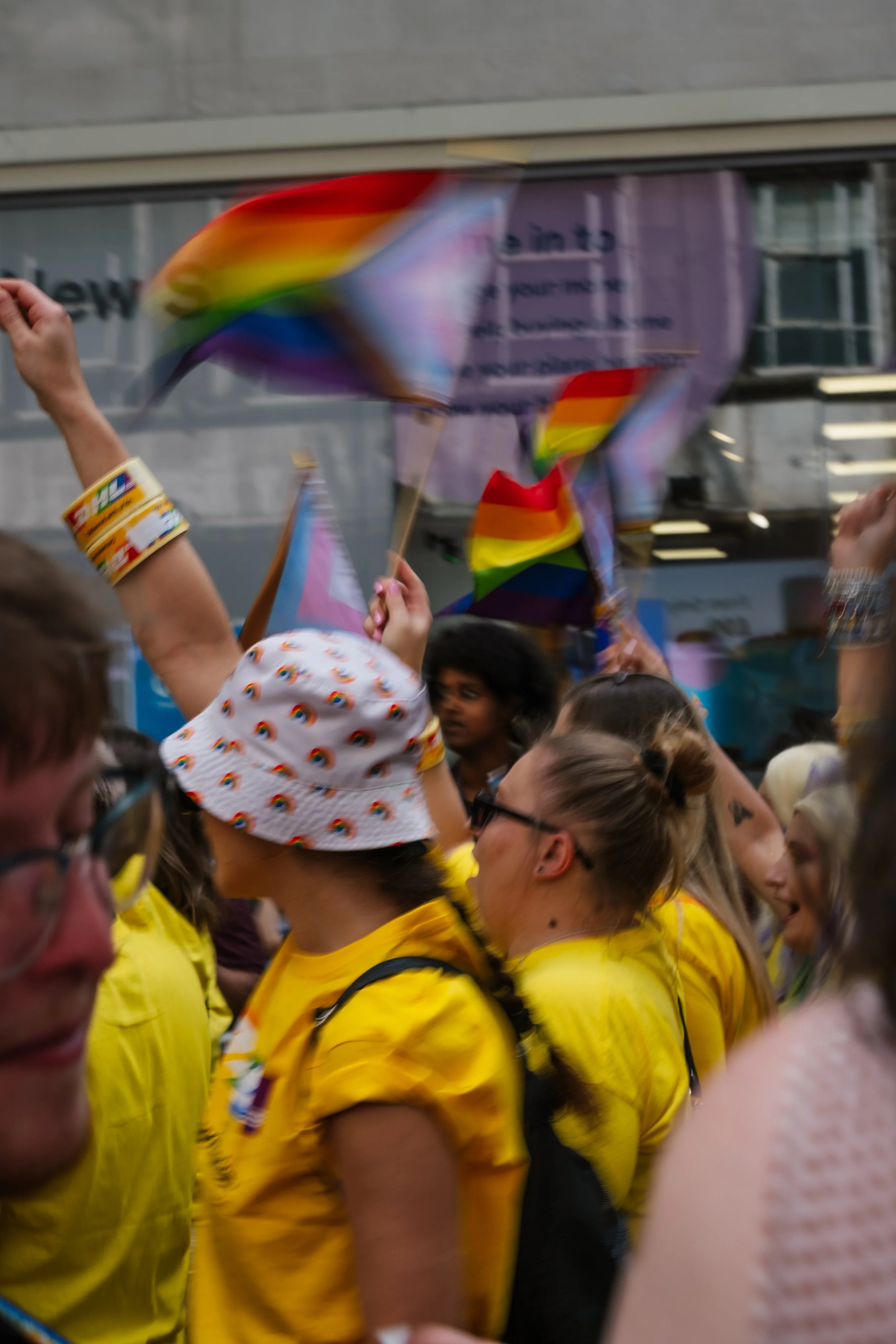 People gathered at an indoor event, many wearing yellow shirts and holding rainbow pride flags.