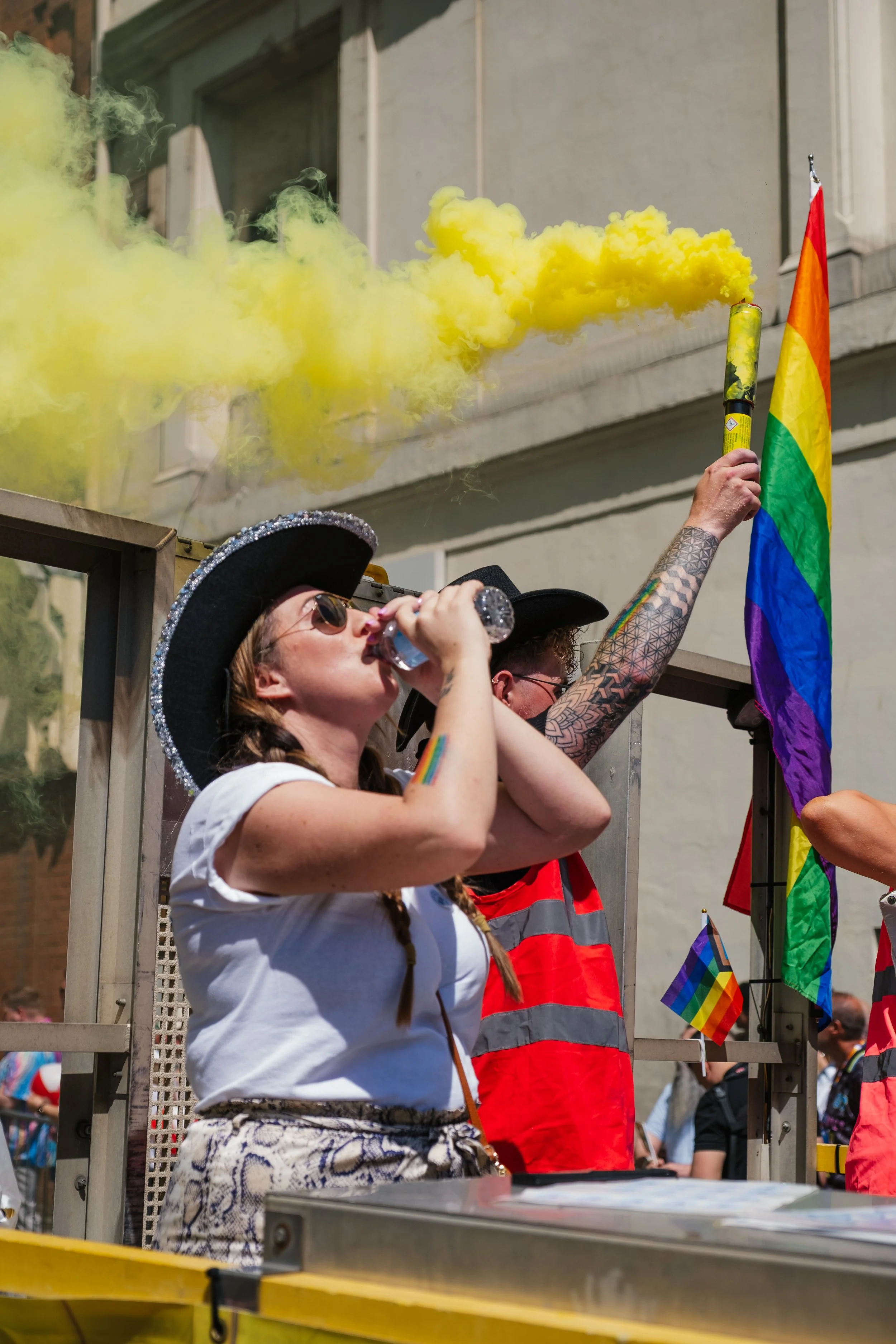 A woman wearing a black hat with silver rhinestones and sunglasses, blowing yellow smoke from a smoke grenade during a pride parade. She is also wearing a white T-shirt and patterned shorts, with a rainbow Pride wristband on her arm. In the backgroun