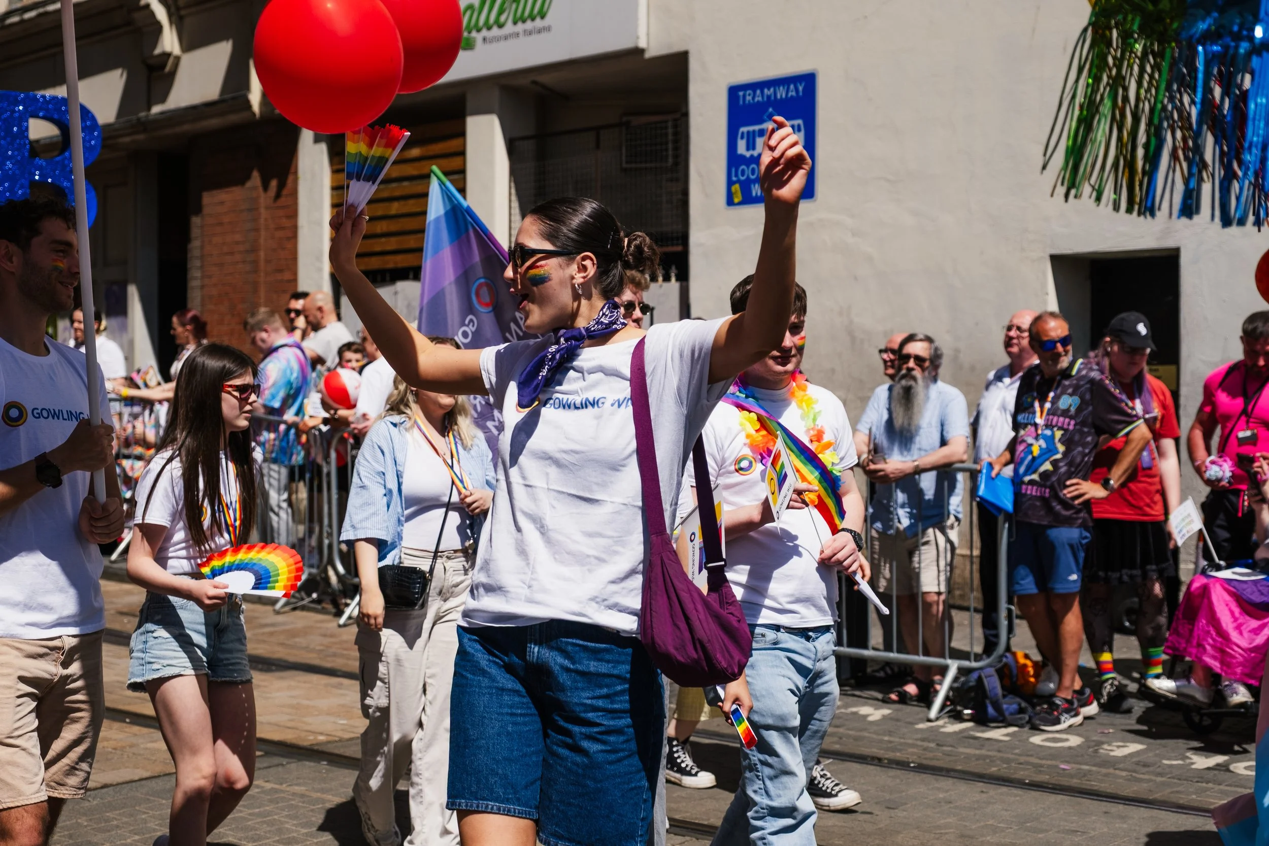 People participating in a pride parade or festival, wearing rainbow-themed accessories and holding rainbow flags and balloons, with a crowd of spectators in the background.