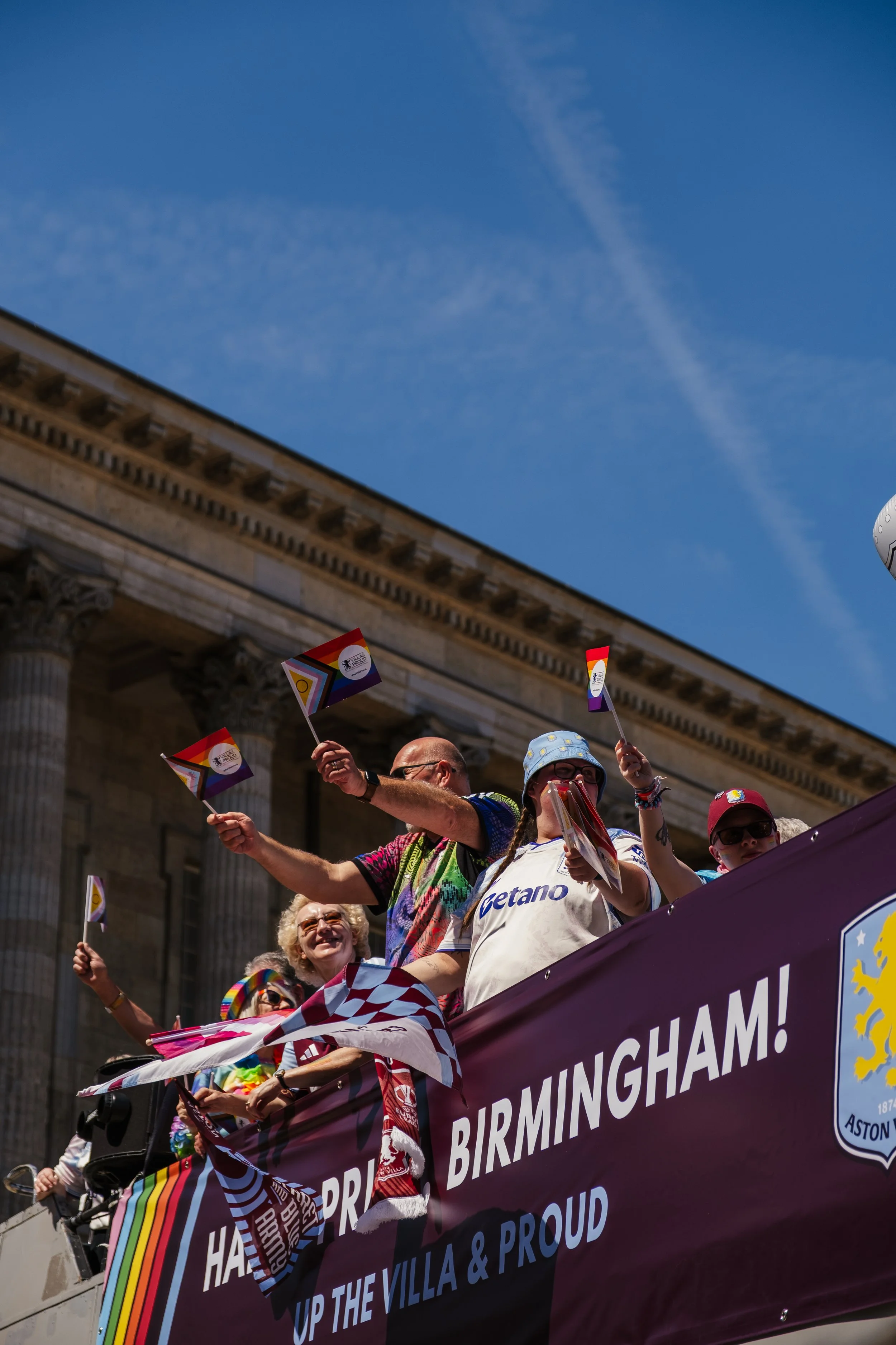 People participating in a pride parade, holding rainbow flags and a large banner that reads 'UP THE VILLA & PROUD.' The parade is taking place in front of a historic building with classical columns under a clear blue sky.