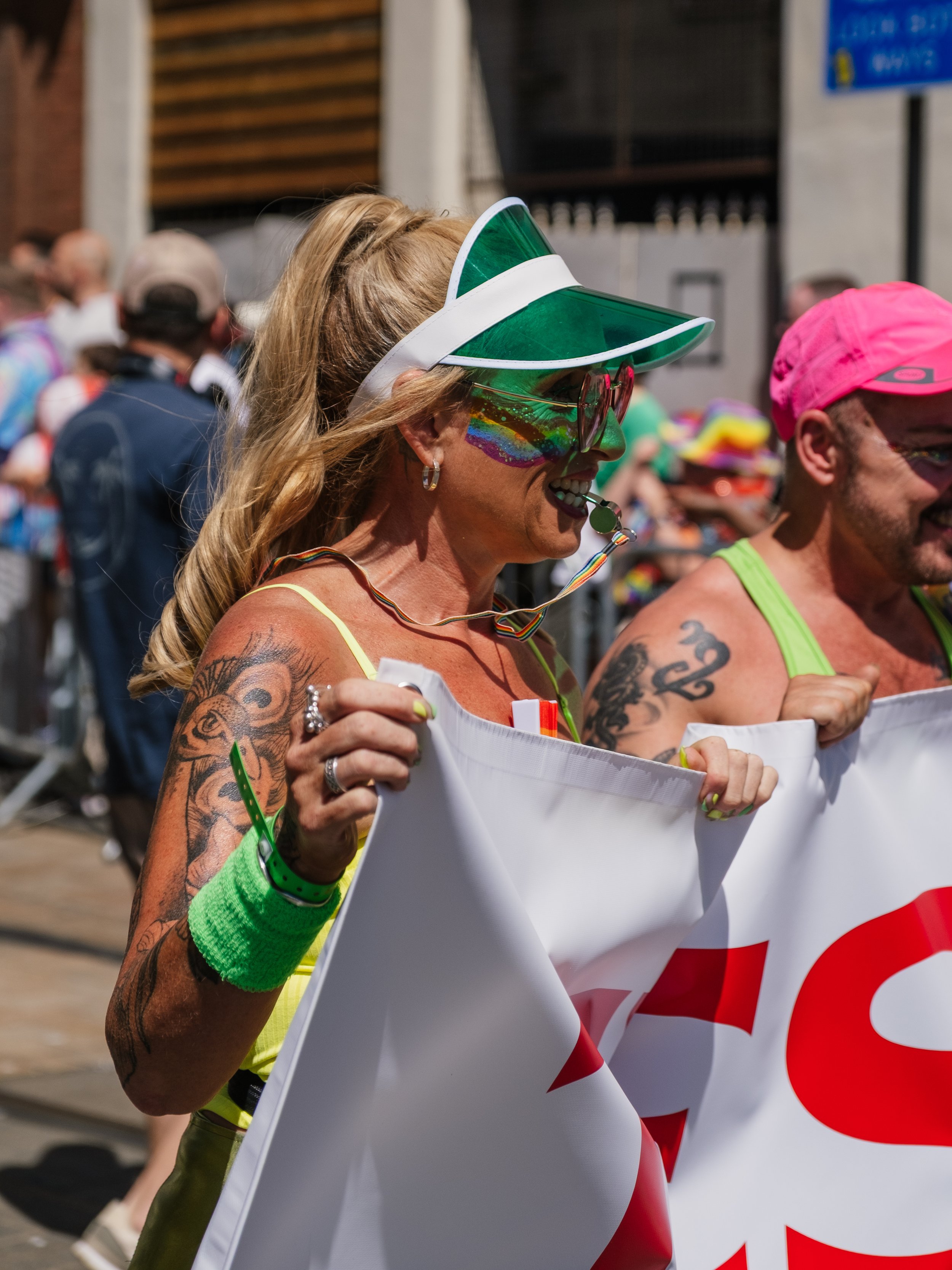 A woman with tattoos, rainbow face paint, wearing a rainbow visor and sunglasses, holding a banner at a pride event.