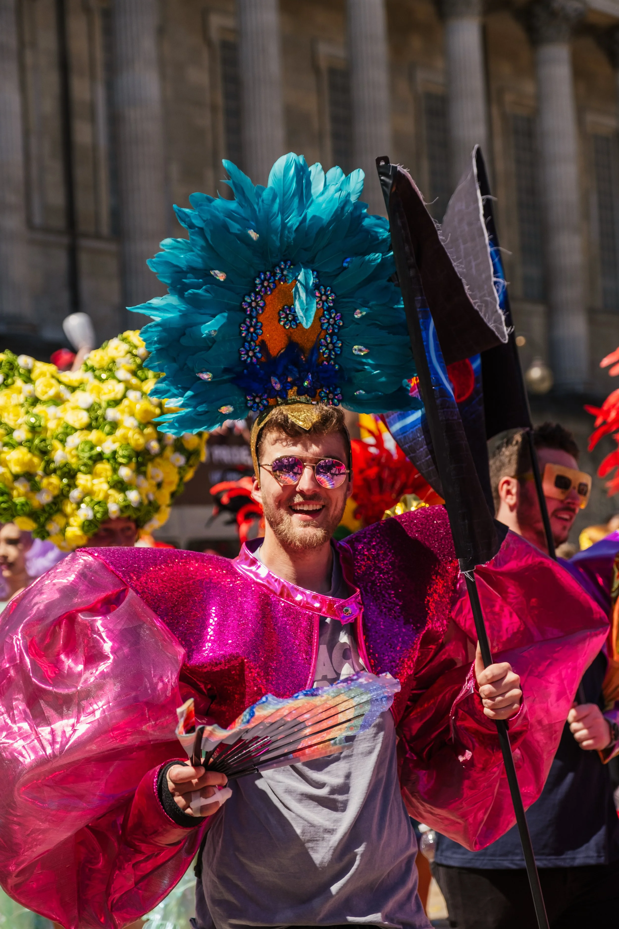 Person wearing a large blue feathered headdress decorated with colorful beads, pink reflective sunglasses, a shiny pink and purple costume, and holding a fan, participating in a parade or celebration with other individuals in colorful outfits.