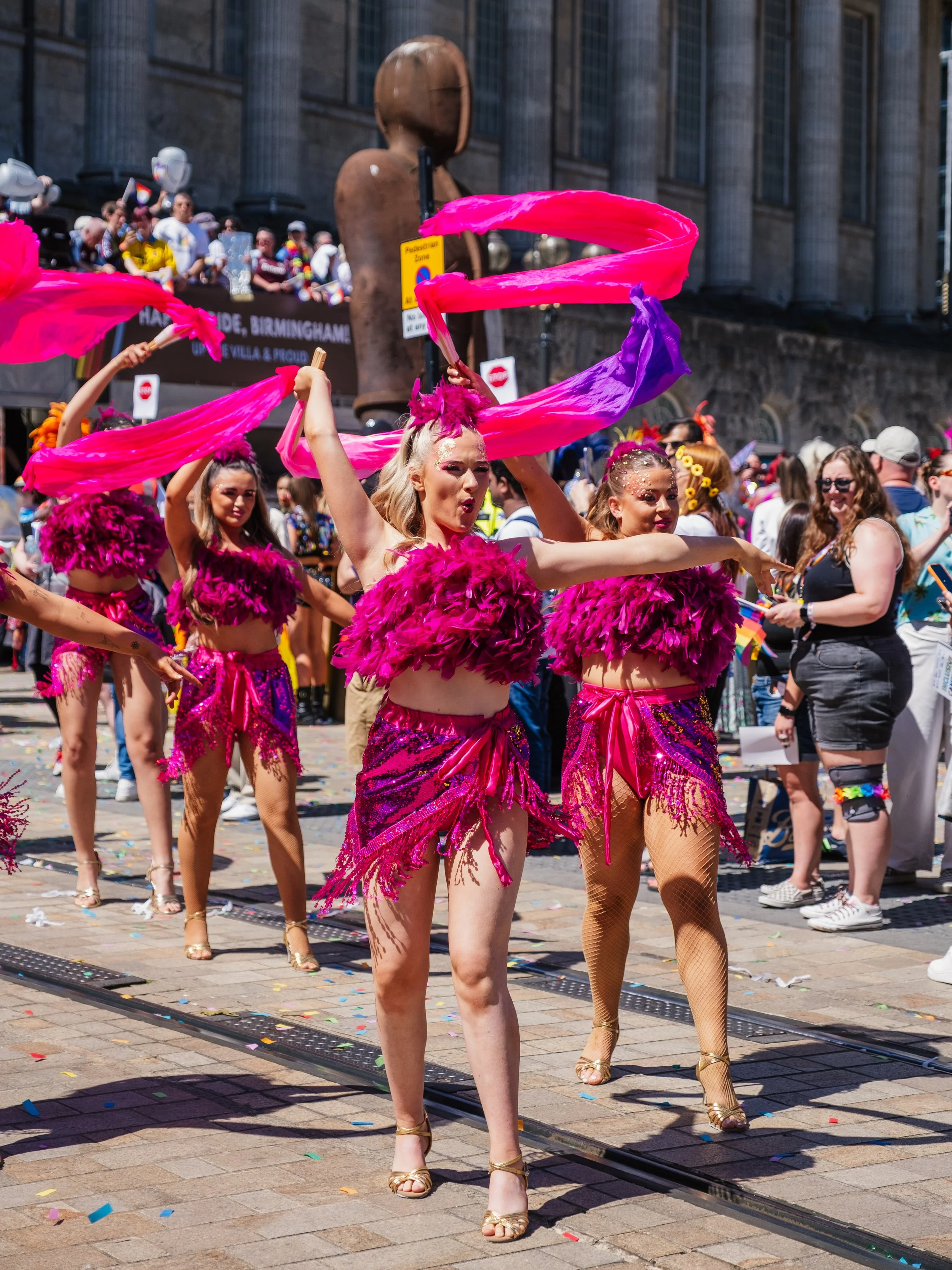 Group of dancers in colorful costumes performing during a parade on a city street, with a large crowd and buildings in the background.
