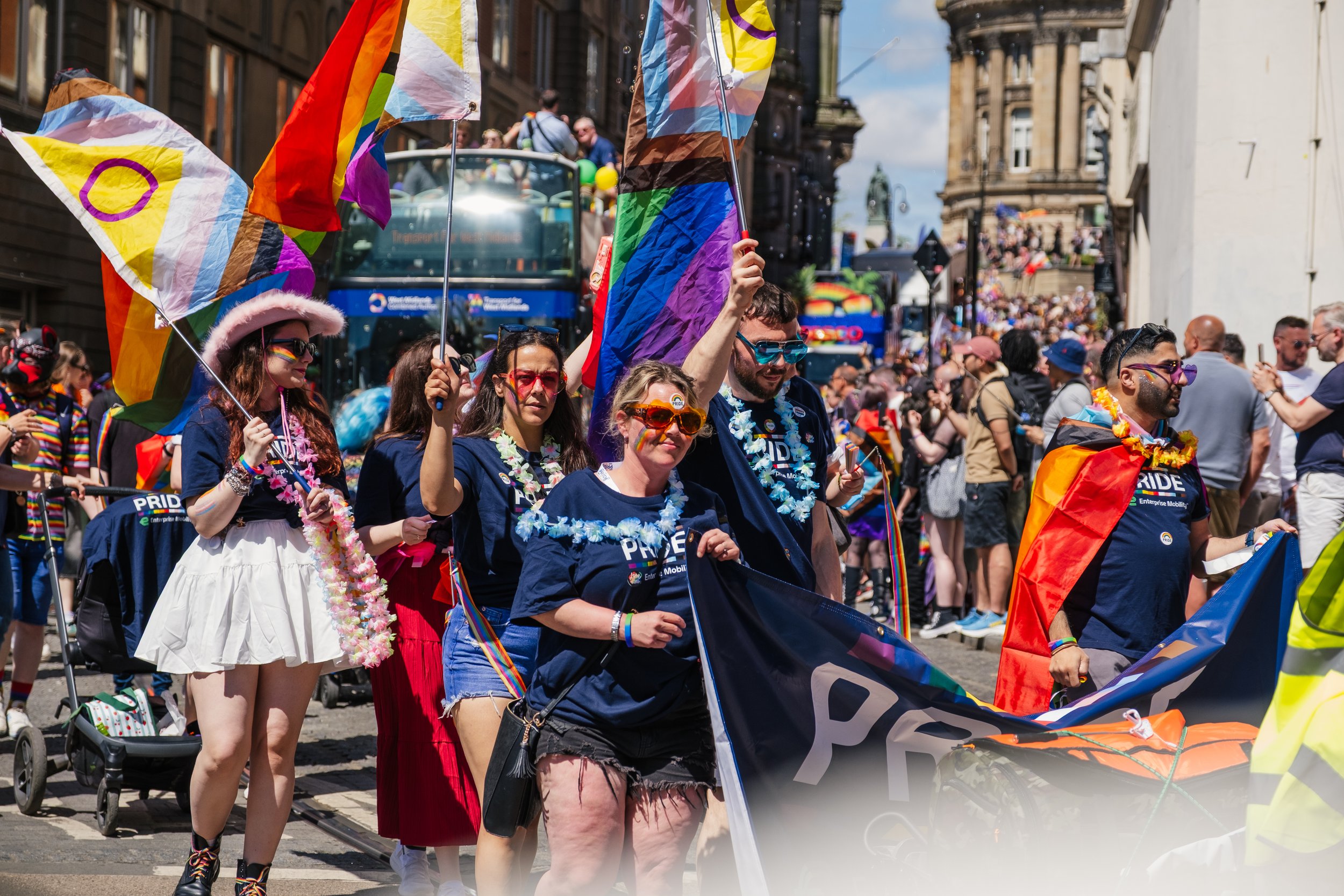 People participating in a pride parade holding rainbow flags and wearing colorful accessories in a city street with onlookers in the background.