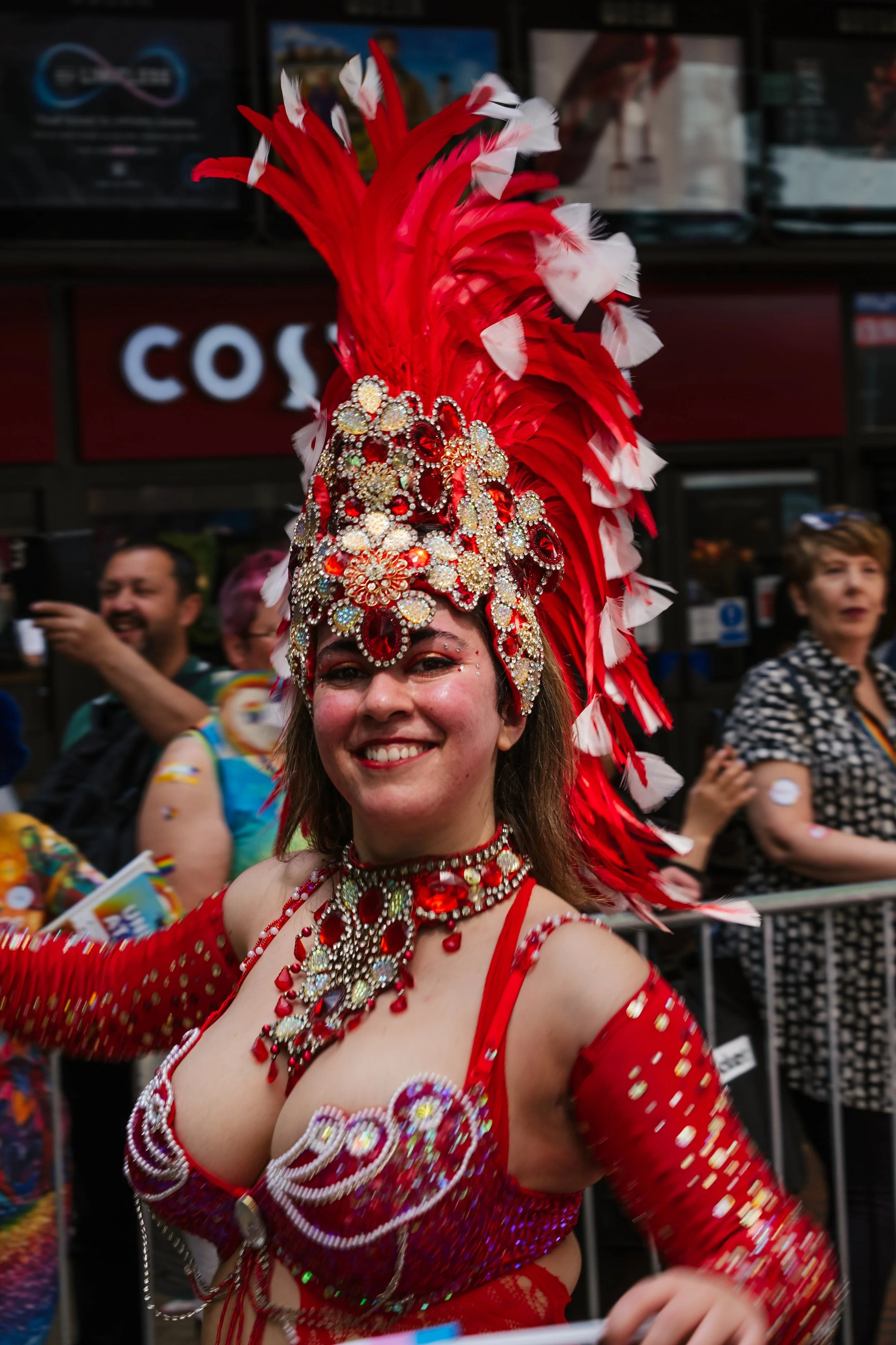 A woman smiling and dressed in a red, bejeweled carnival costume with a large feathered headdress, participating in a parade or festival with onlookers in the background.
