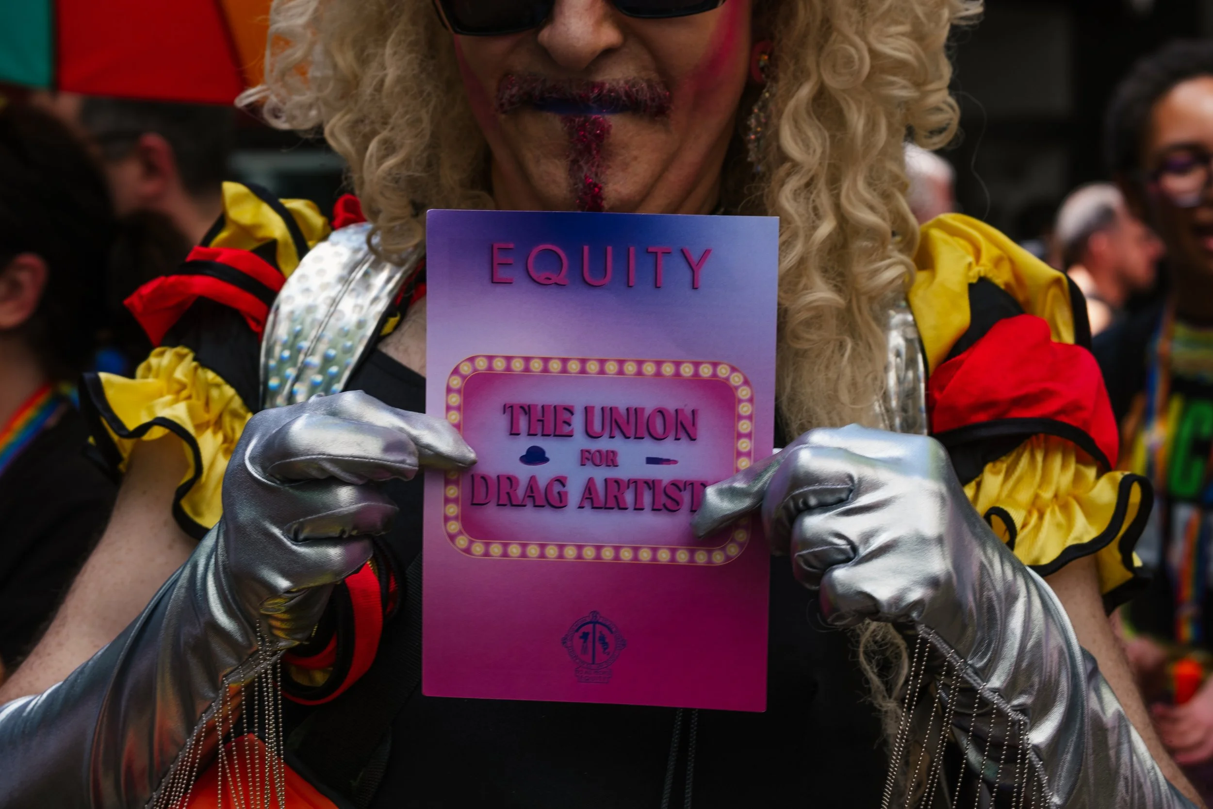 Person dressed as a drag artist holding a pink sign that reads 'EQUITY THE UNION FOR DRAG ARTISTS' at a pride event, with other attendees visible in the background.