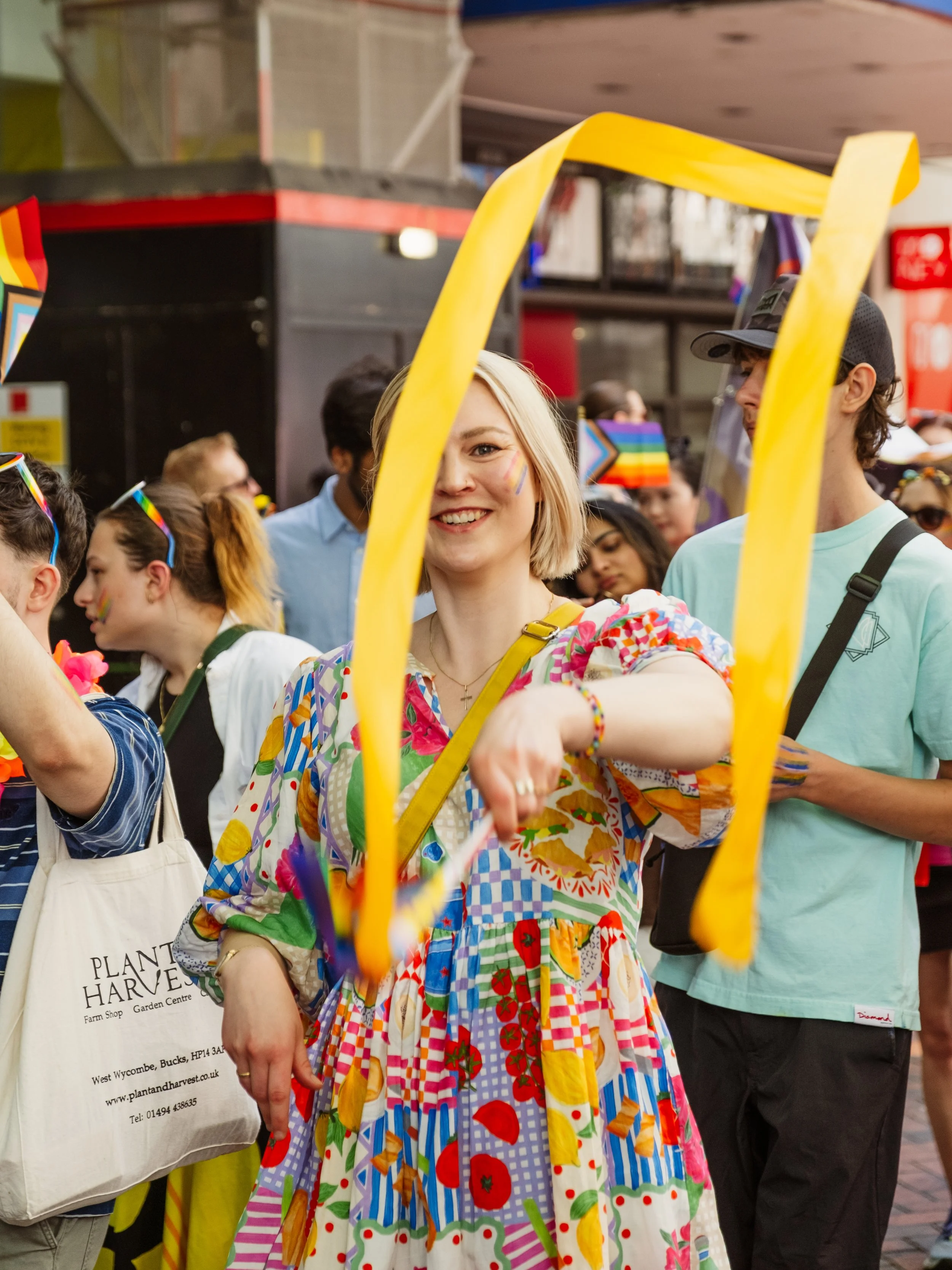 A woman with blonde hair smiling, wearing a colorful patchwork dress, at a pride event with people in the background holding rainbow flags and wearing rainbow accessories.