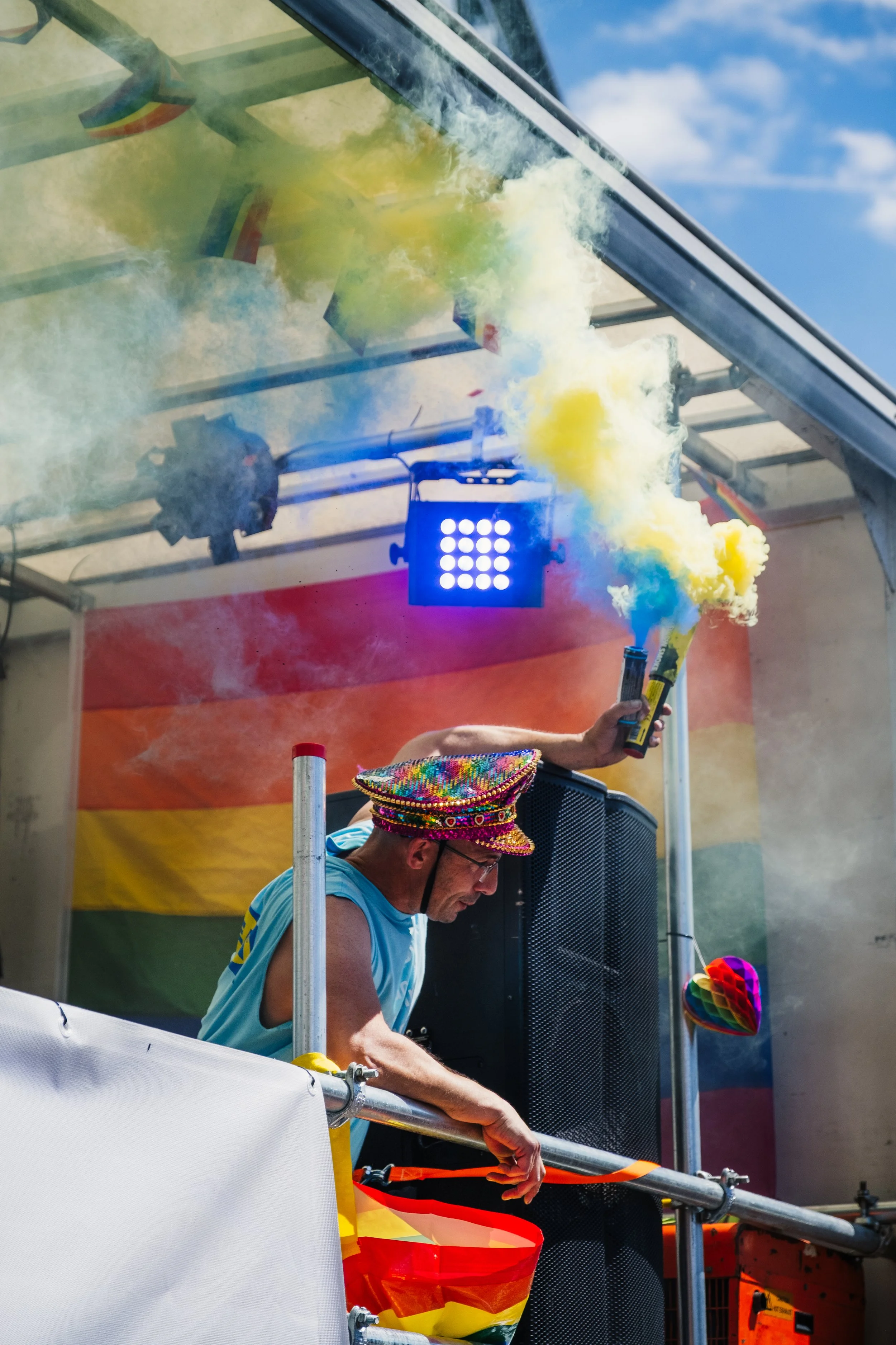 A man wearing a colorful rainbow hat and blue tank top is DJing at an outdoor pride event. Smoke and colorful flags are visible, with blue sky and white clouds in the background.