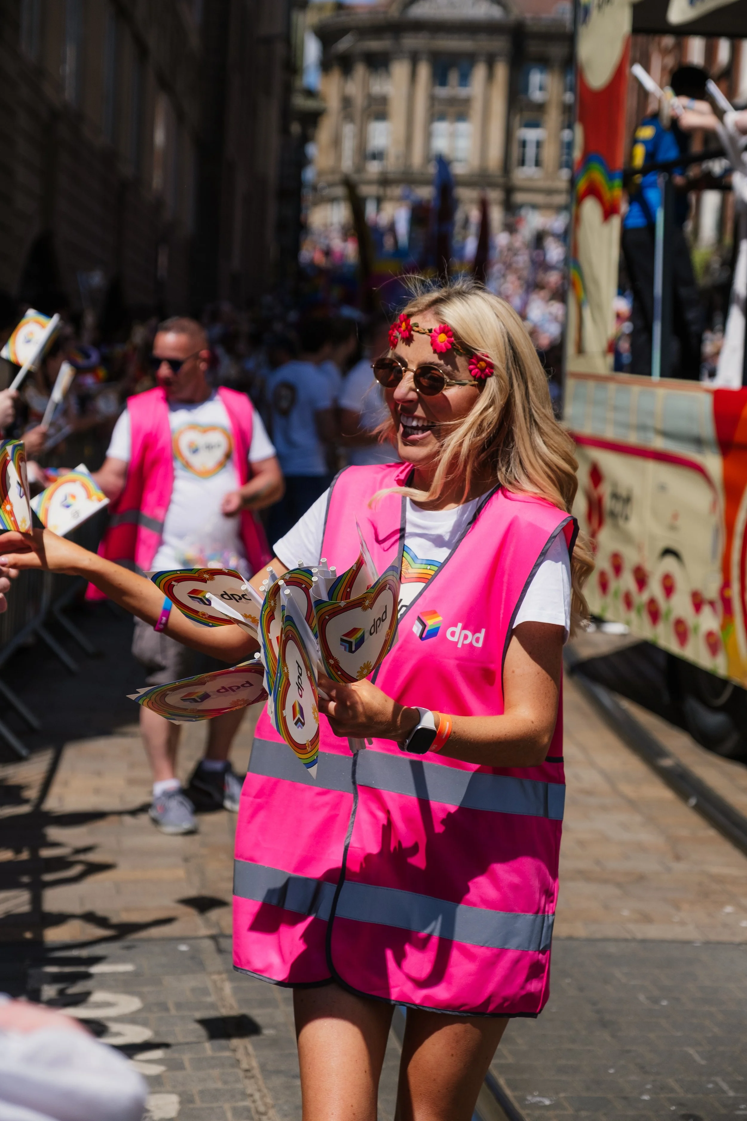 A woman with long blonde hair wearing sunglasses, a pink safety vest, and a white T-shirt is smiling and handing out rainbow-themed pamphlets during a parade. She is wearing a flower headband and a wristwatch. There are others dressed similarly, and 