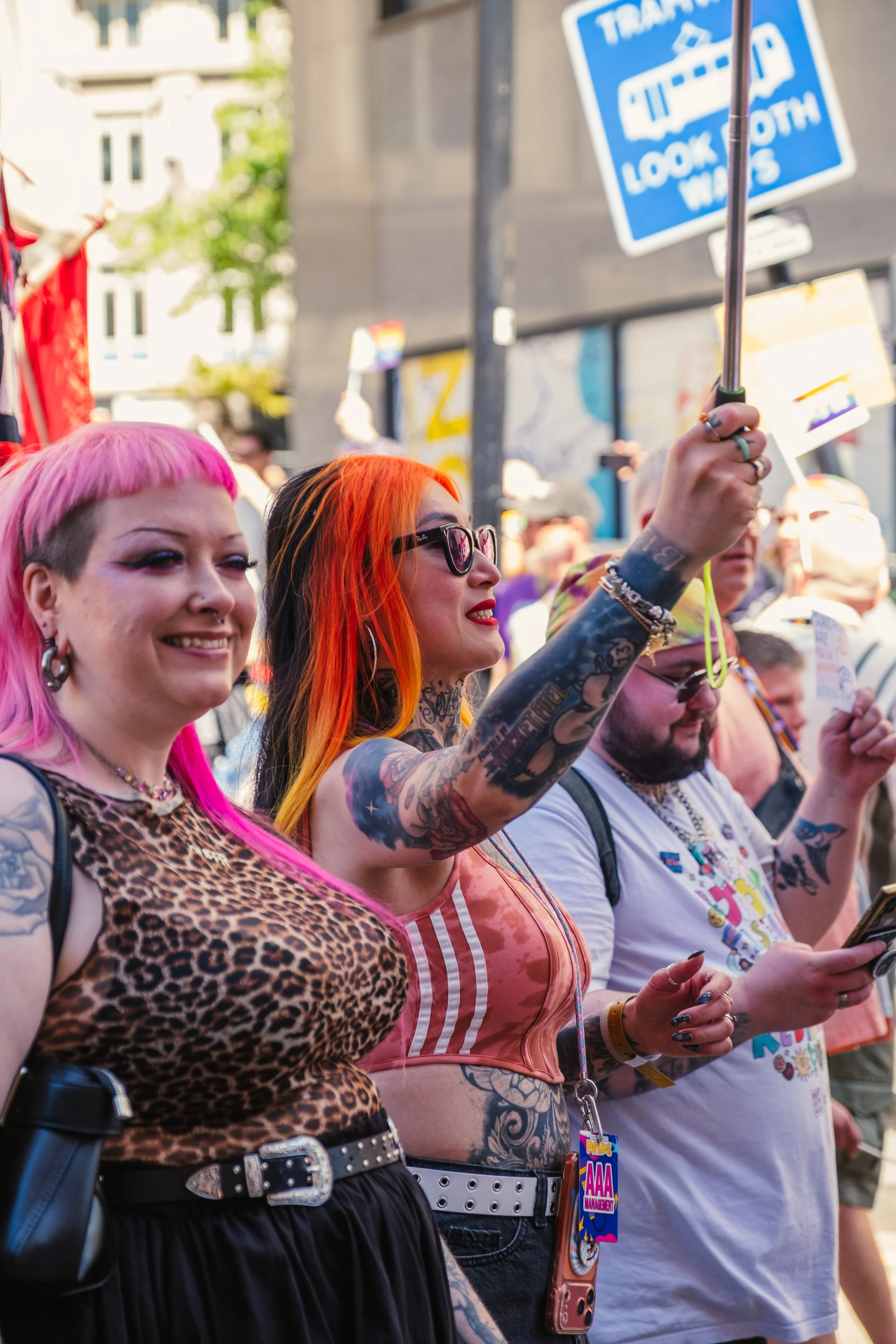 People at a parade or protest, with a focus on two women in the foreground. One woman has pink hair and a leopard print top, the other has orange hair, glasses, and tattoos, holding a rainbow flag on a stick.