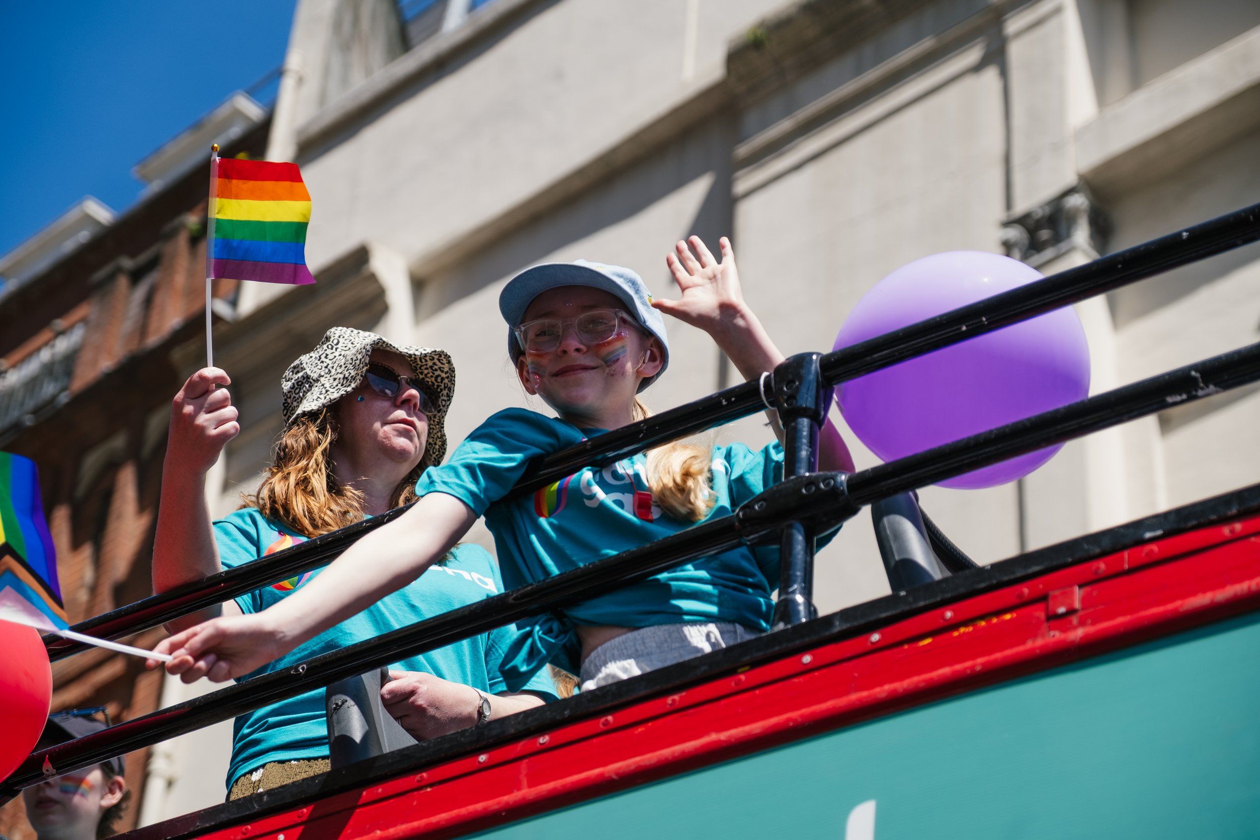 People riding on a float during a parade, with rainbow Pride flags and balloons.