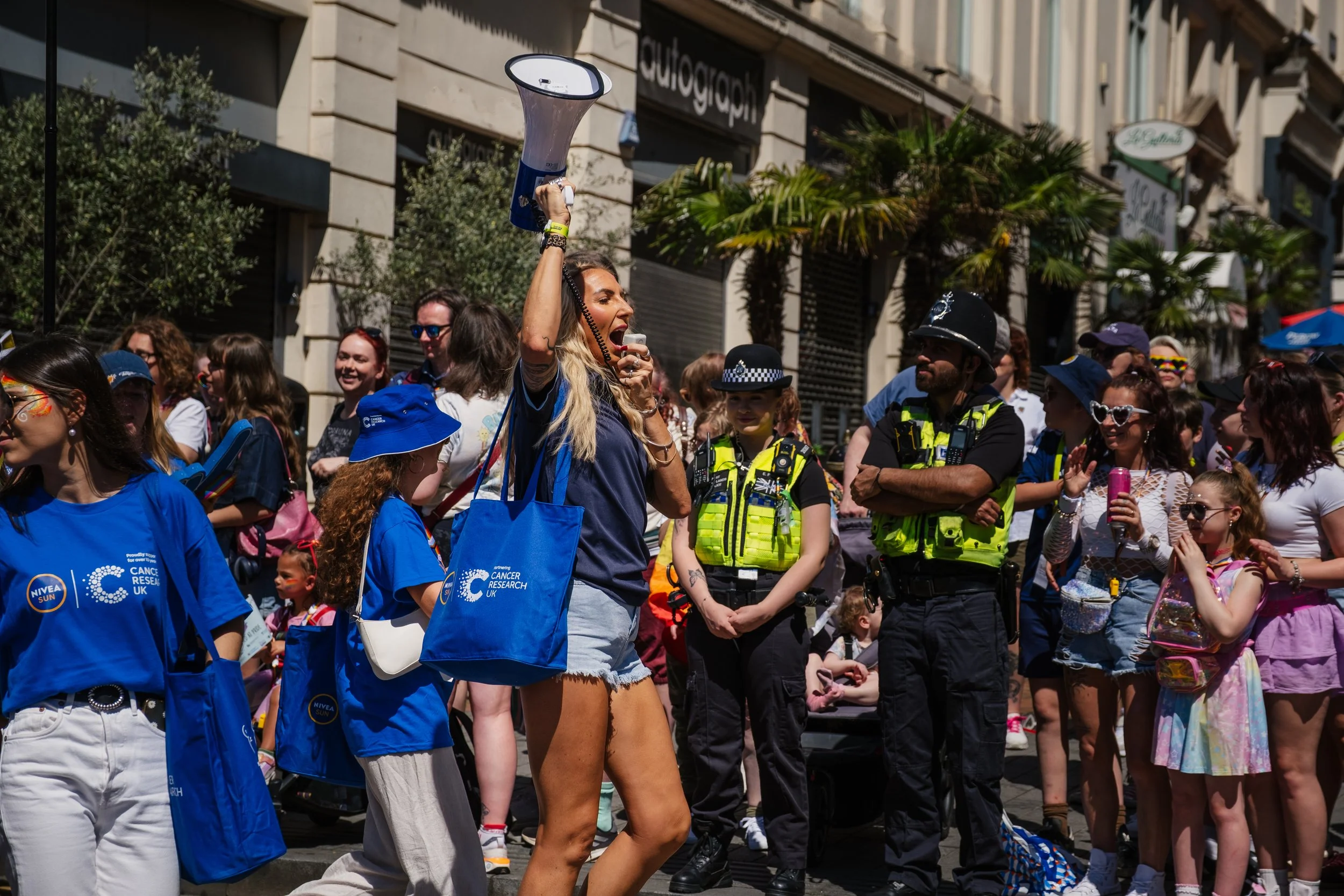 A woman speaking into a megaphone at a public event or protest, surrounded by a crowd including police officers and various participants.