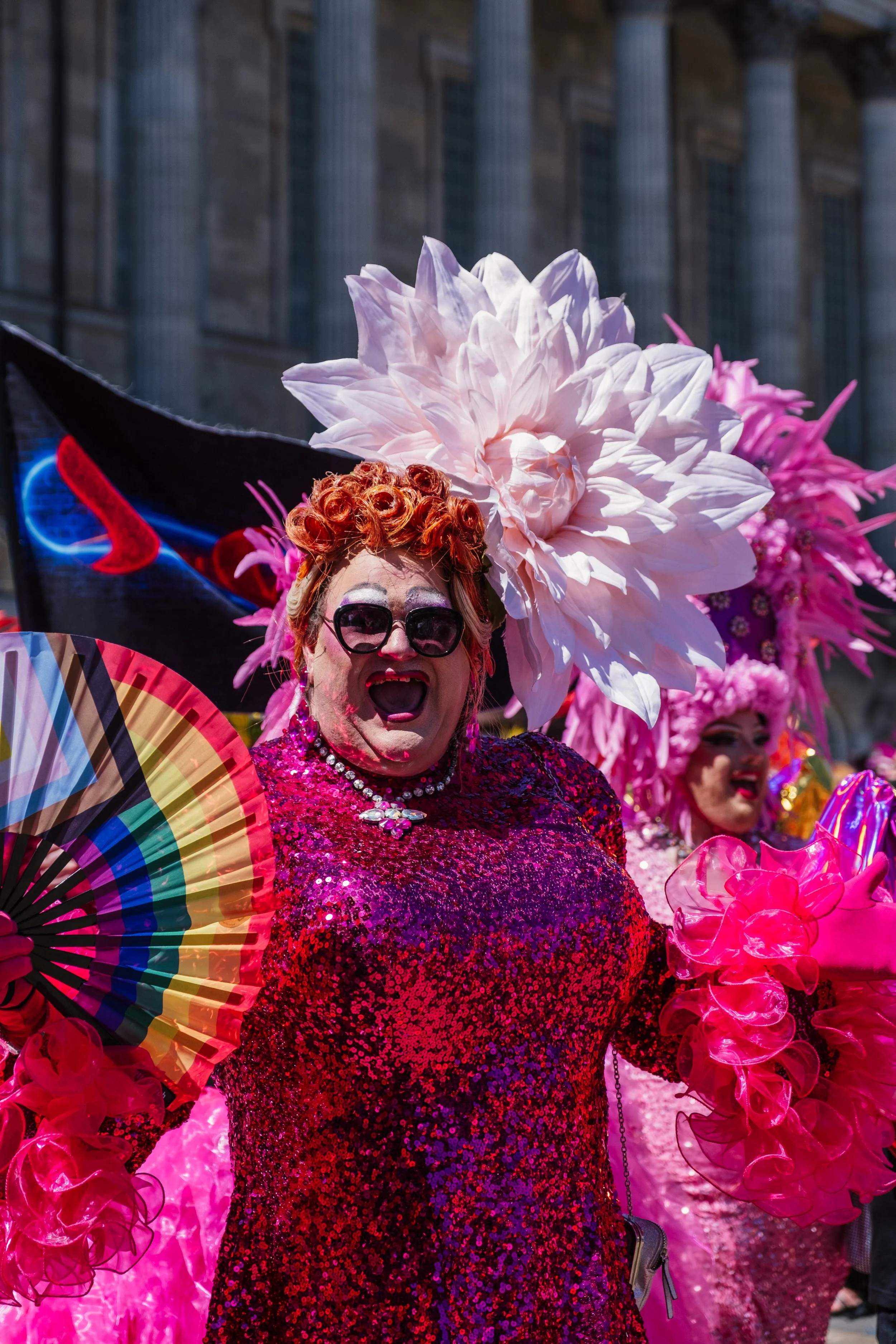 Person in a colorful sequined dress wearing large floral headpiece and sunglasses, holding a rainbow-colored fan during a parade or celebration.