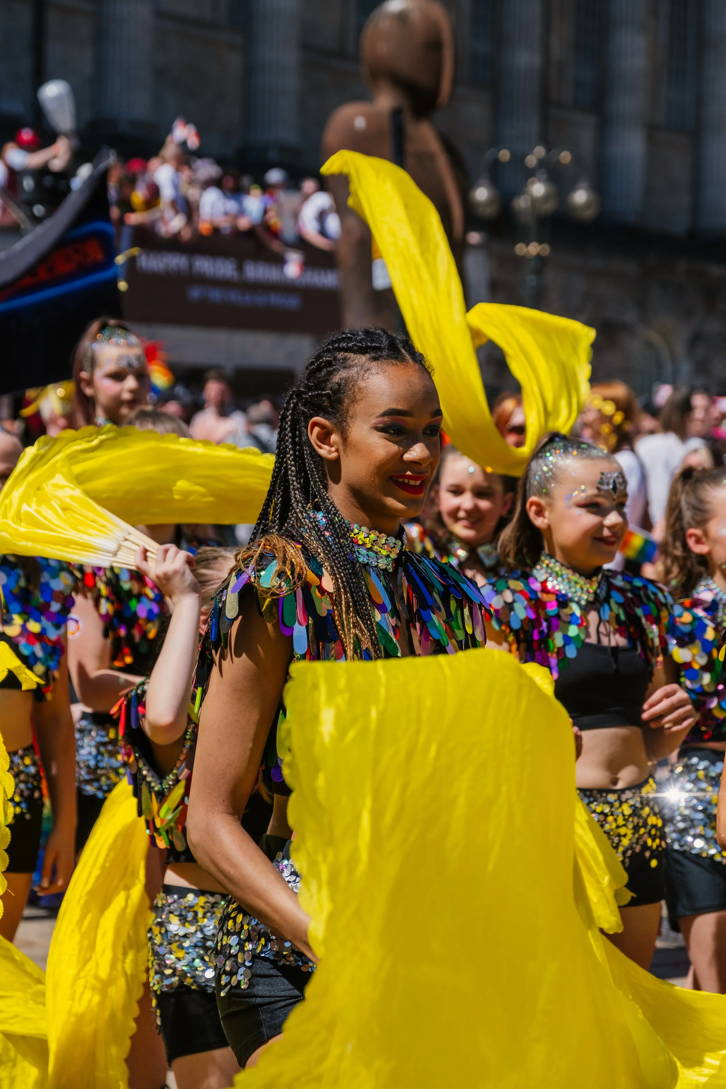 A group of young women wearing colorful, sequined costumes with large yellow fabric wings, participating in a parade or festival, surrounded by a crowd of spectators.