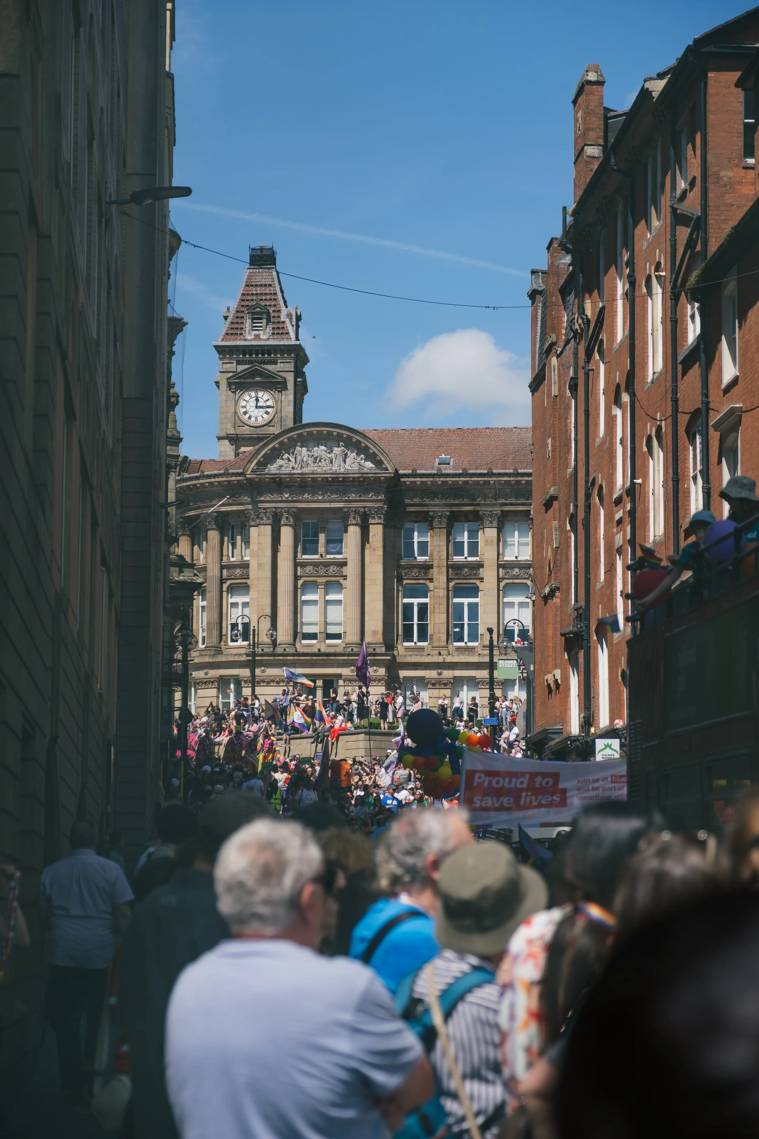 Street scene with a crowd gathering for a pride parade, with rainbow flags and colorful balloons. A historic building with a clock tower is visible in the background under a clear blue sky.