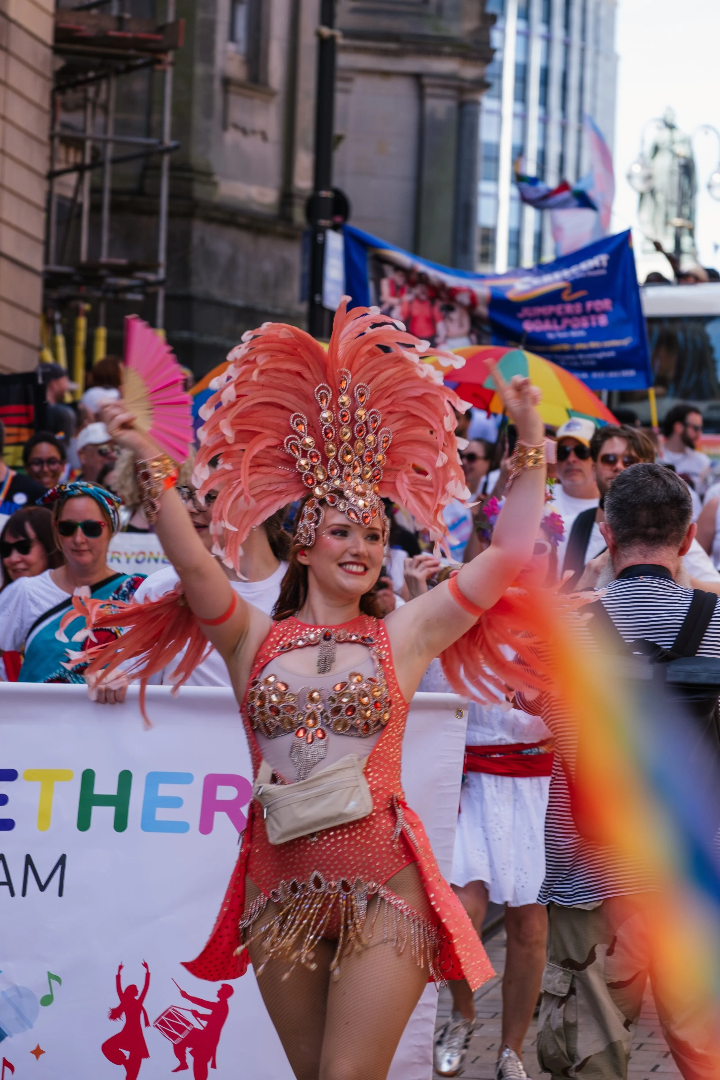 Performer in orange costume with feathered headdress dancing during a parade, with a crowd and banners in the background.