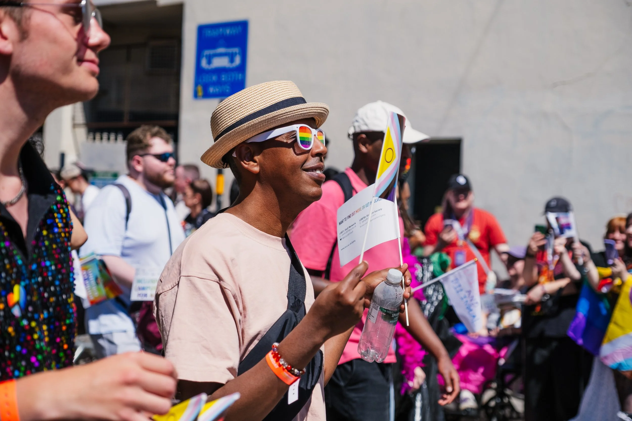 A group of diverse people participating in a pride parade, with some wearing rainbow-themed accessories and sunglasses, holding water bottles and signs, walking in a lively outdoor setting on a sunny day.