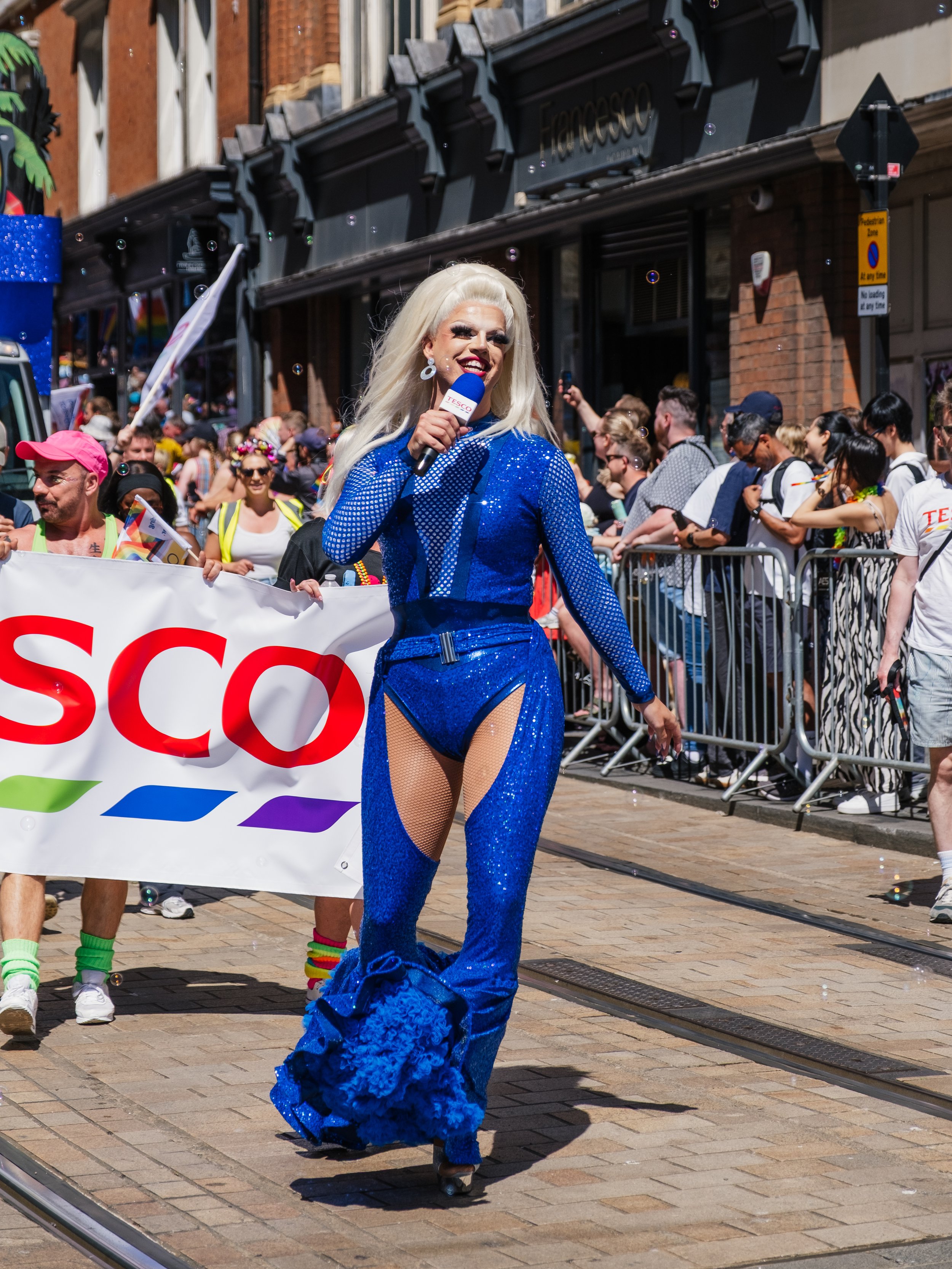 A drag queen with platinum blonde hair and glamorous makeup, dressed in a sparkling blue outfit with fishnet tights, roller skating in a parade while holding a microphone with the Tesco logo, behind a crowd of spectators.