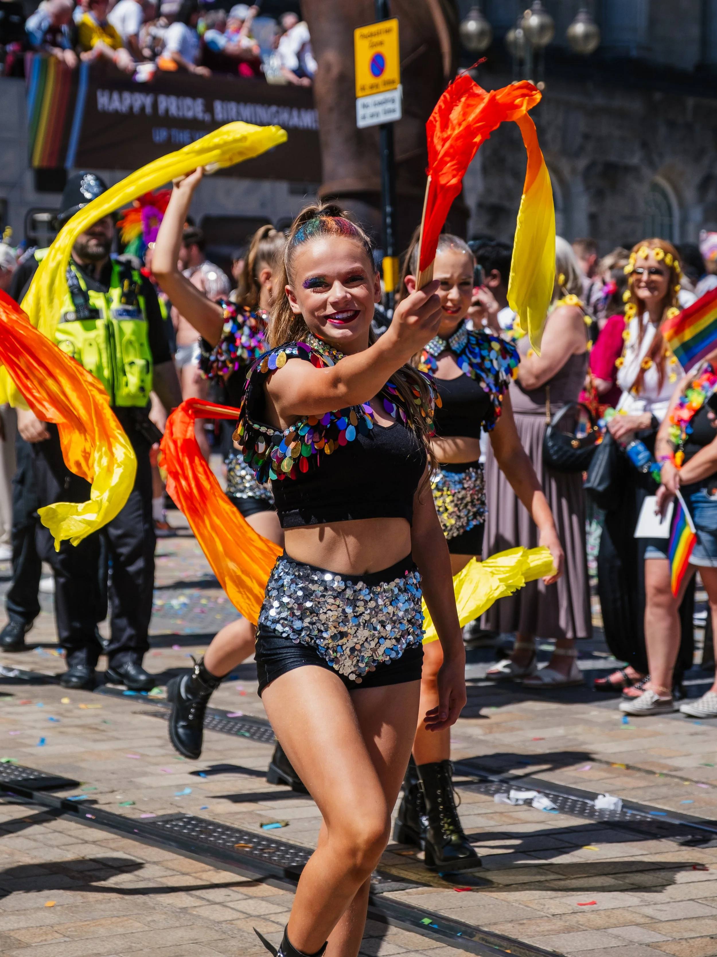 A woman in a sequined black crop top and matching shorts dances with colorful flowing scarves at a pride parade. She is smiling and surrounded by other participants and spectators, with a sign and a crowd in the background.